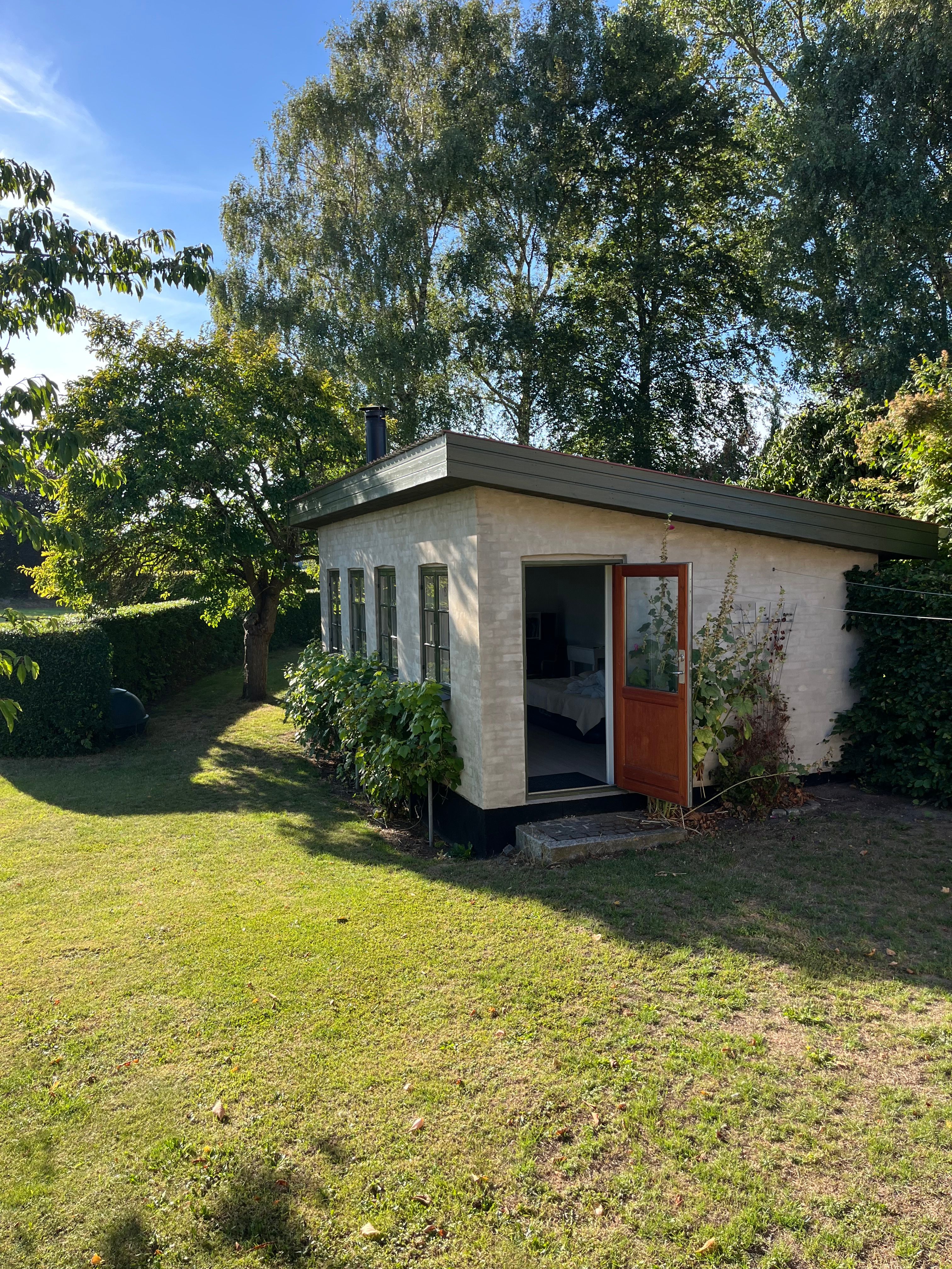 White-painted brick garden studio with an open wooden door and casement windows, set on a lawn among trees