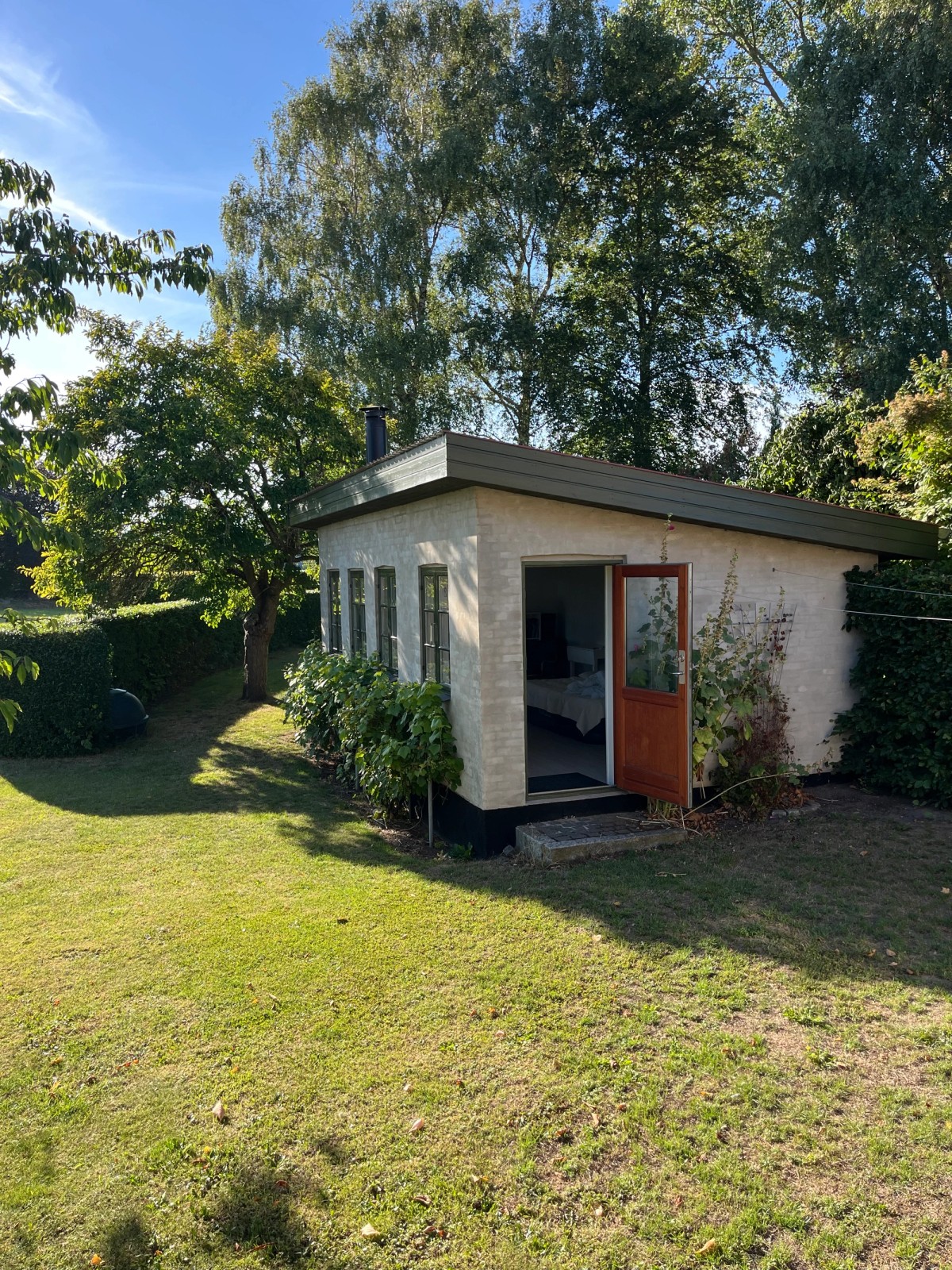 White-painted brick garden studio with an open wooden door and casement windows, set on a lawn among trees