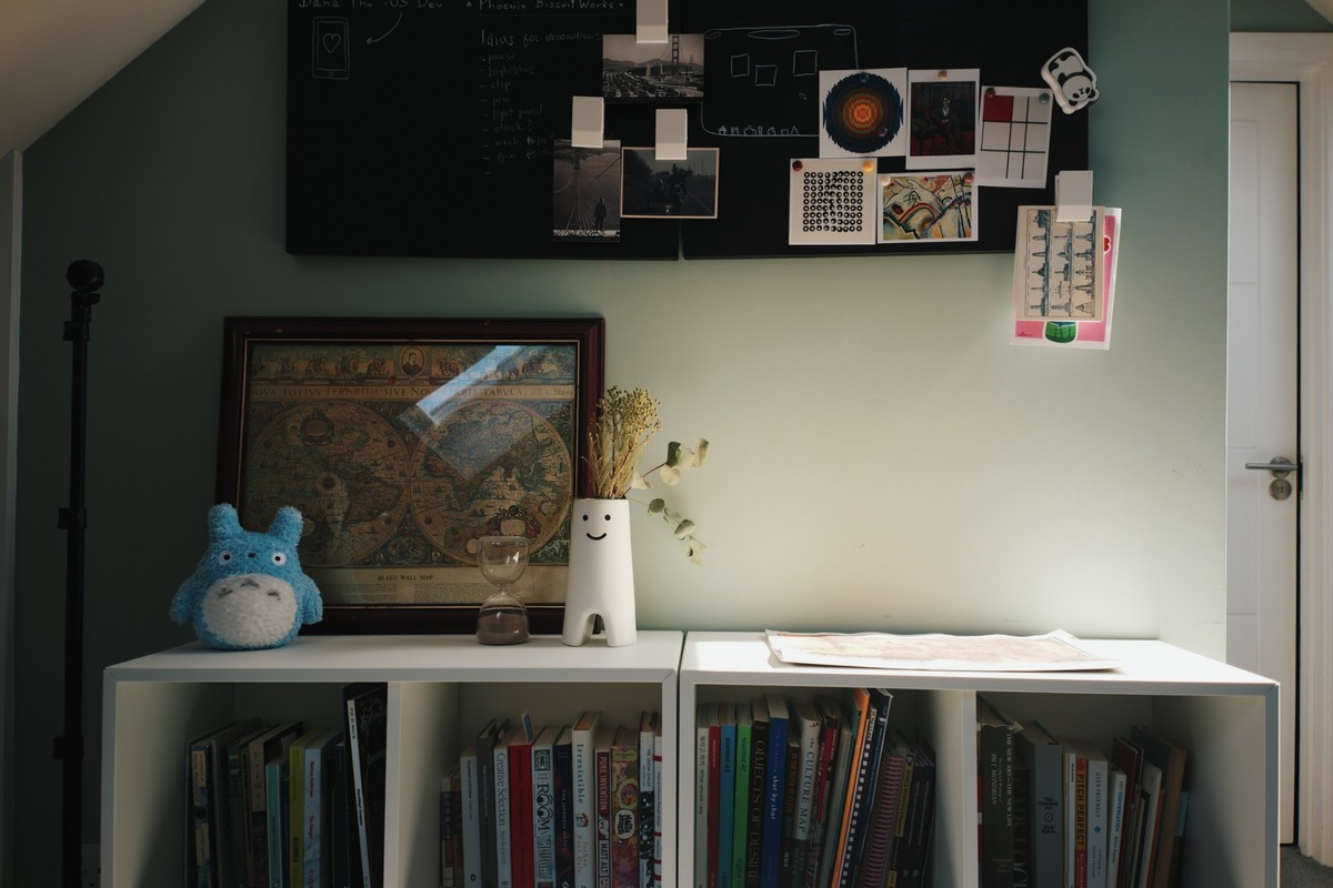 The far wall of the office: a black board pinned with photographs and art prints above a white bookcase holding books, a framed antique world map and a smiling vase.