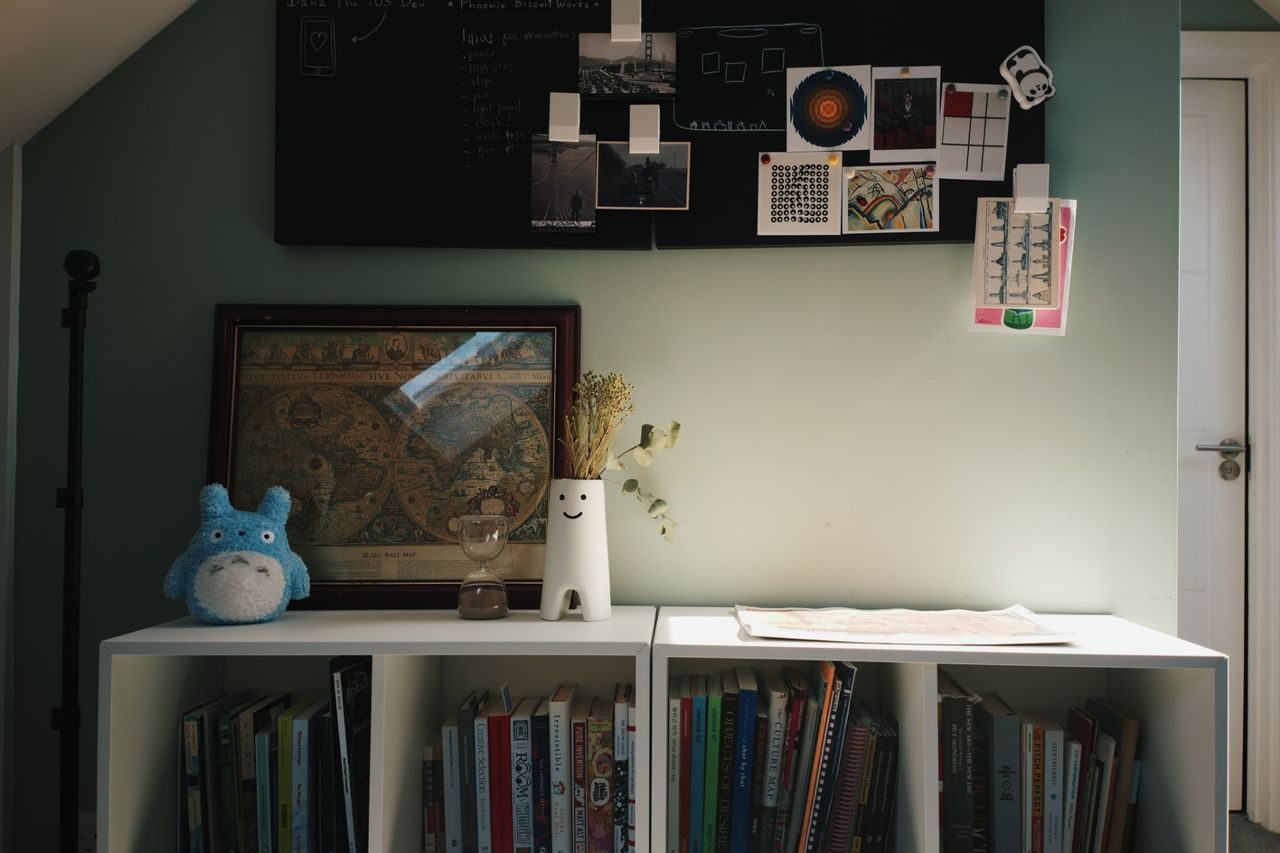 The far wall of the office: a black board pinned with photographs and art prints above a white bookcase holding books, a framed antique world map and a smiling vase.