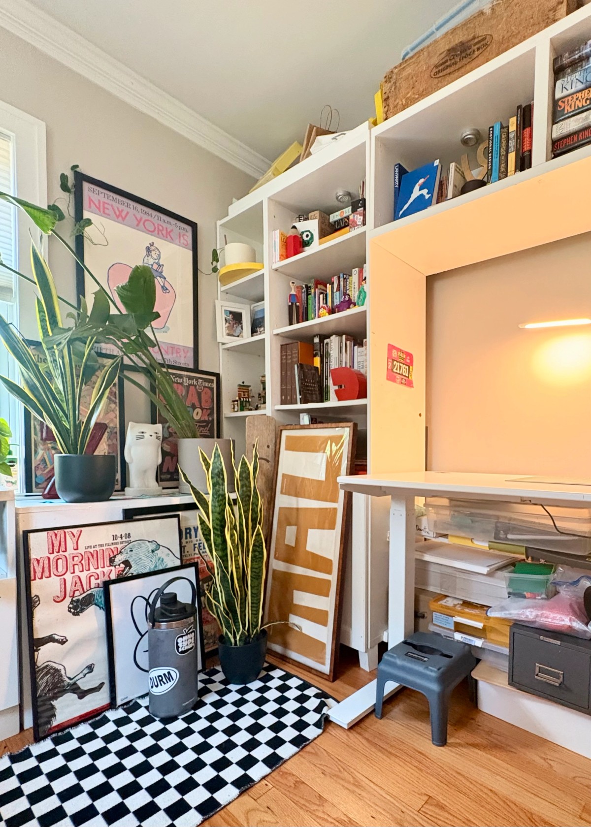 White cube shelves packed with books and design objects beside leaning gig and art posters, snake plants, and a peach-painted alcove, over a black-and-white checkerboard rug.