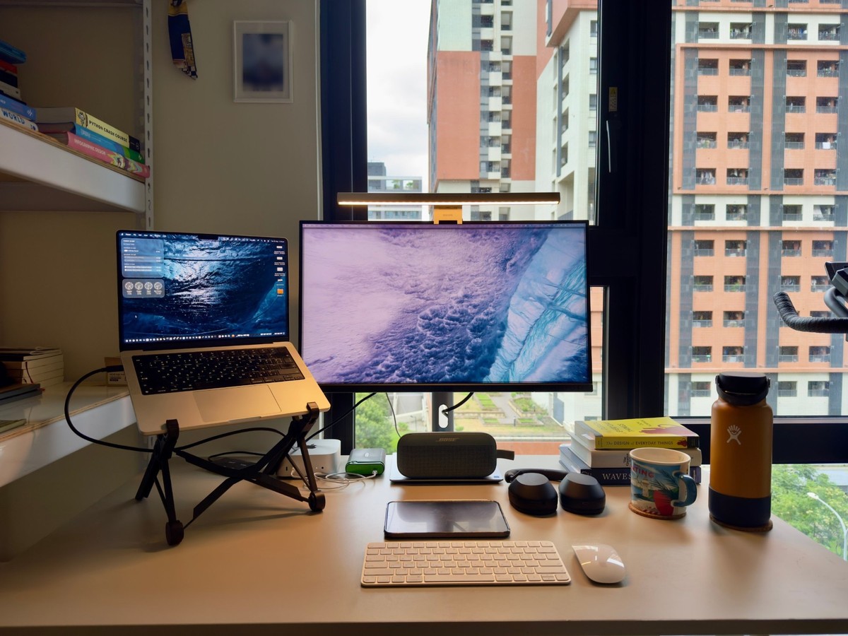 A standing desk in front of a window, with a MacBook Pro on a folding stand, an ASUS monitor lit by a clamp-on light bar, a Bose speaker, headphones, a stack of books, an orange Hydro Flask and a Magic Keyboard and Mouse.