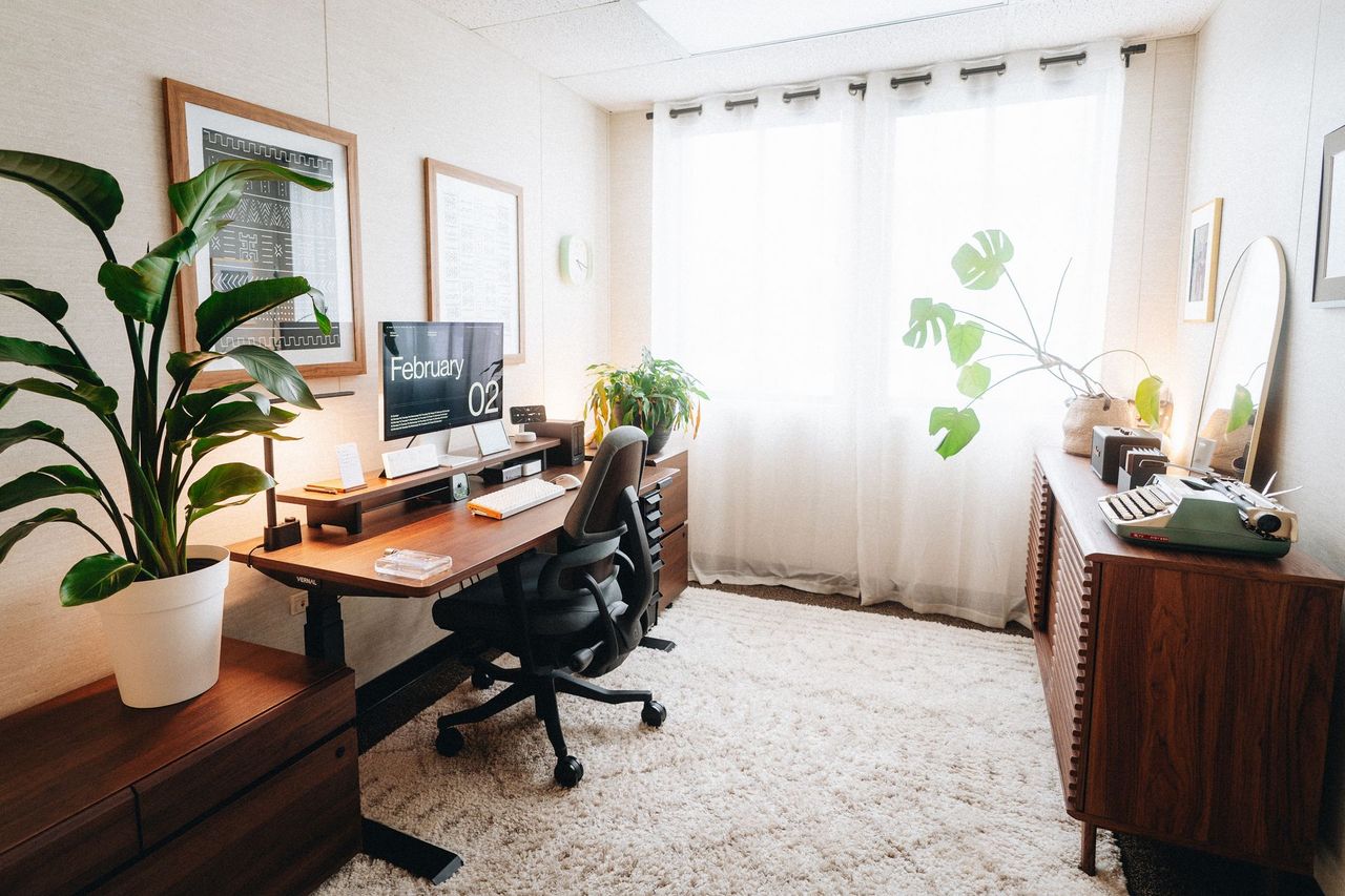 Home office with a wooden standing desk, Apple Studio Display, black office chair, tall plants and sheer white curtains