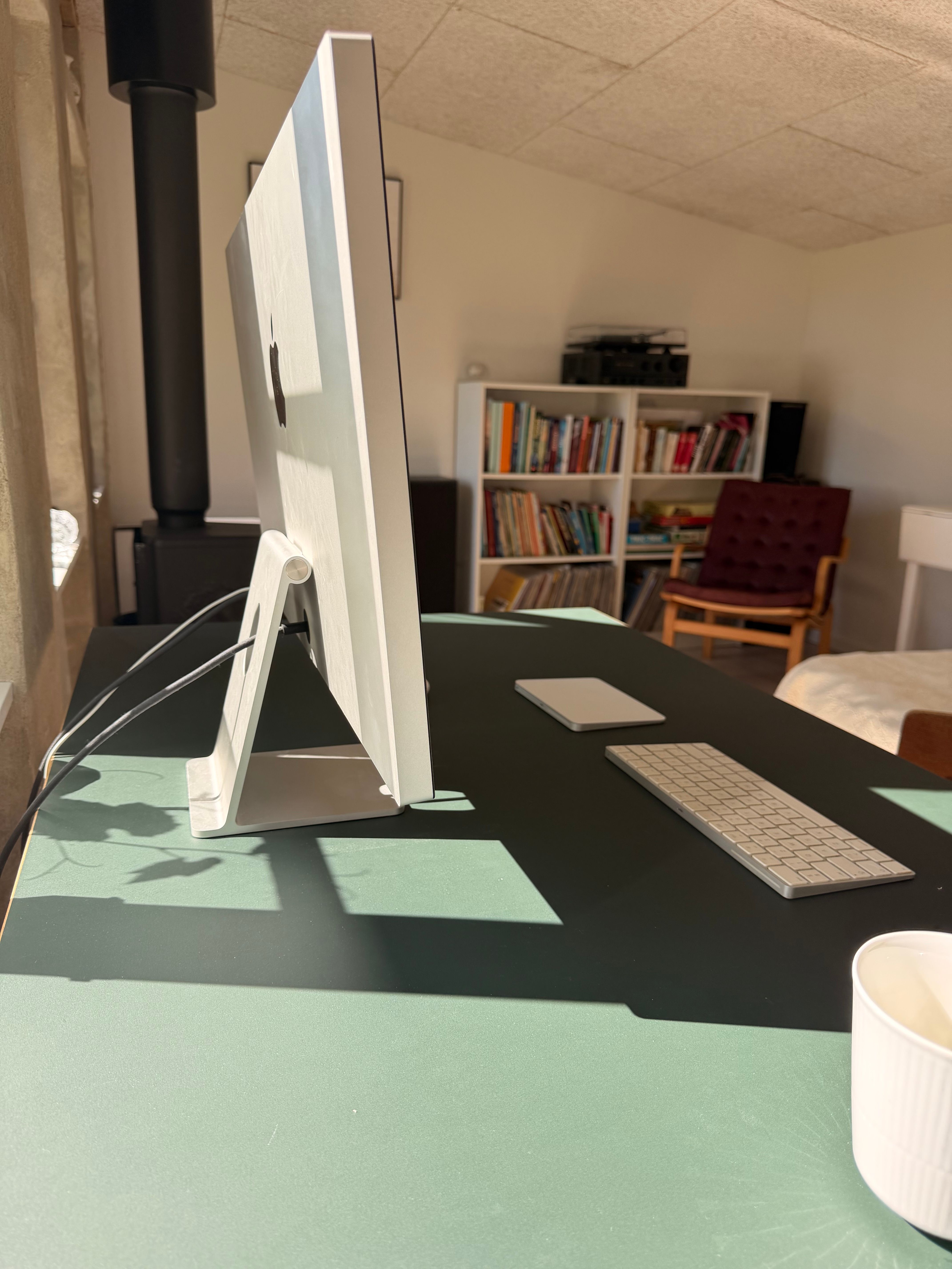 Side view of an Apple Studio Display on an Eiermann table, with a bookshelf, turntable and wood stove behind