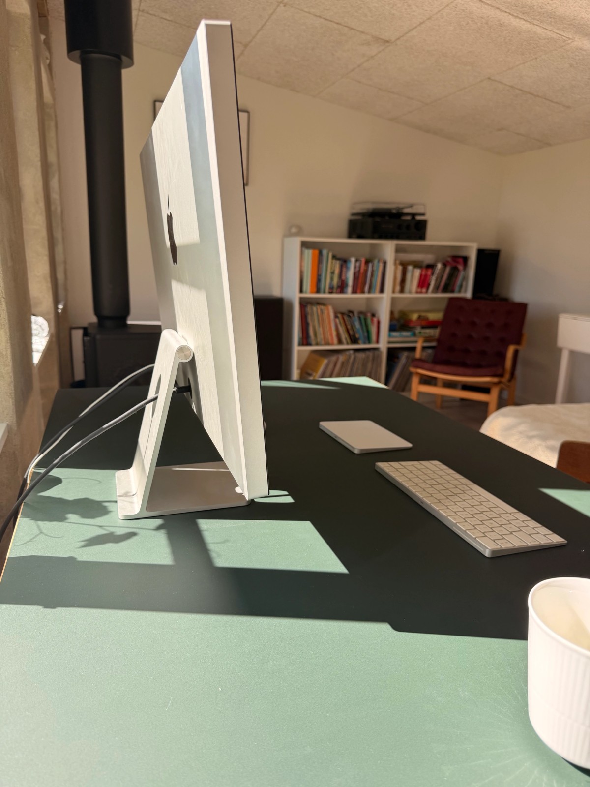 Side view of an Apple Studio Display on an Eiermann table, with a bookshelf, turntable and wood stove behind
