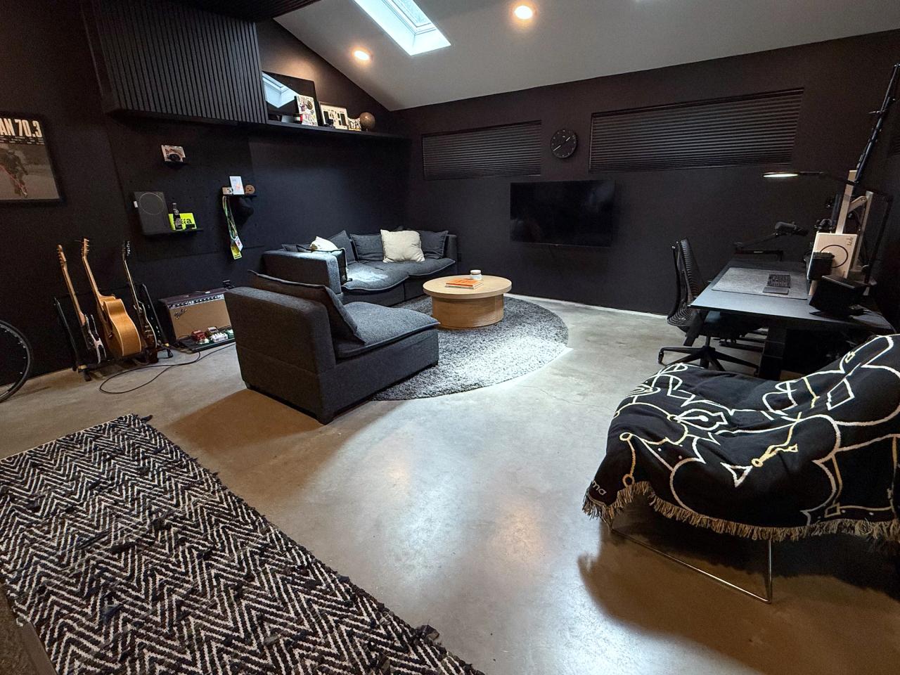 Black-walled studio with skylights, a concrete floor, sofas around a round coffee table, guitars, a wall-mounted TV and a desk on the right