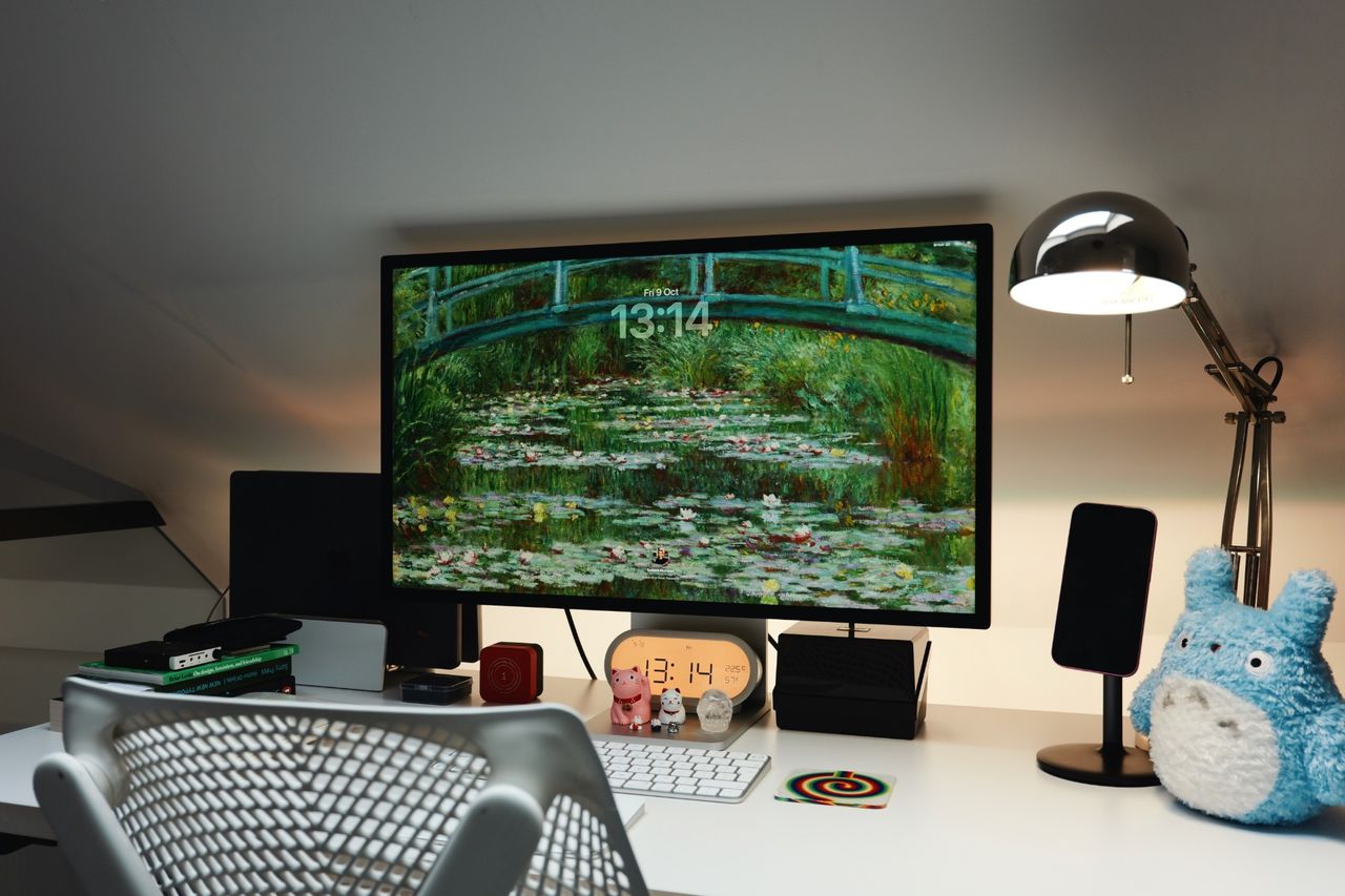 A white desk at night with an Apple Studio Display showing a Monet water lilies lock screen, a chrome anglepoise lamp, a blue Totoro plush and small figures lined up below the monitor.