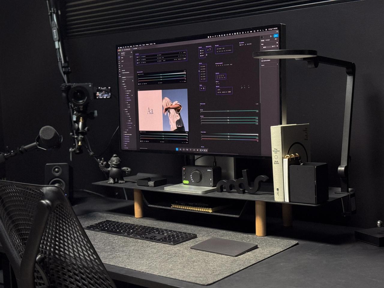 All-black desk setup with an Apple Pro Display XDR on a monitor shelf, keyboard and trackpad on a gray felt mat, a microphone on a boom arm and a mesh chair