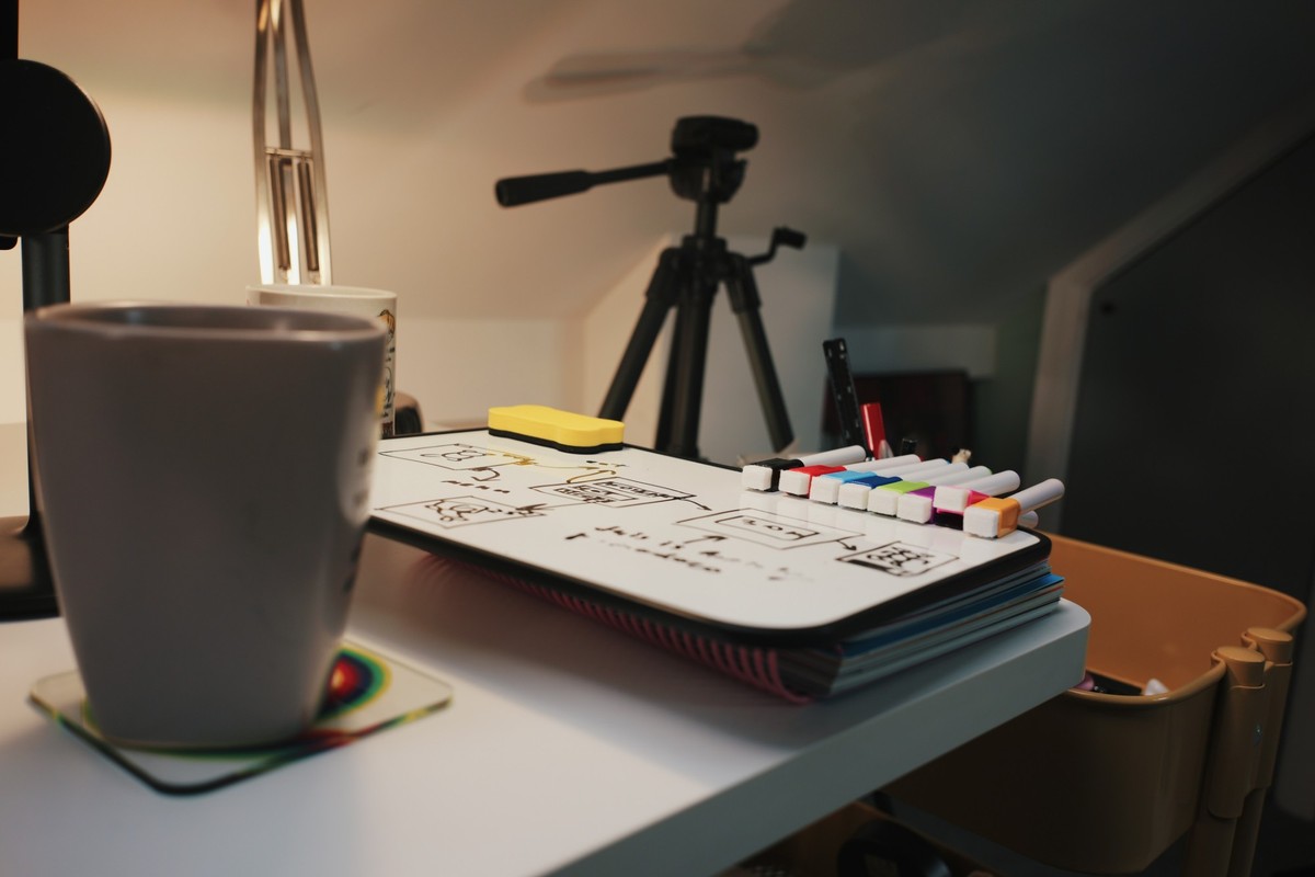A small whiteboard covered in boxy sketches, with colored markers and a yellow eraser resting on it, a mug in the foreground and a tripod behind.