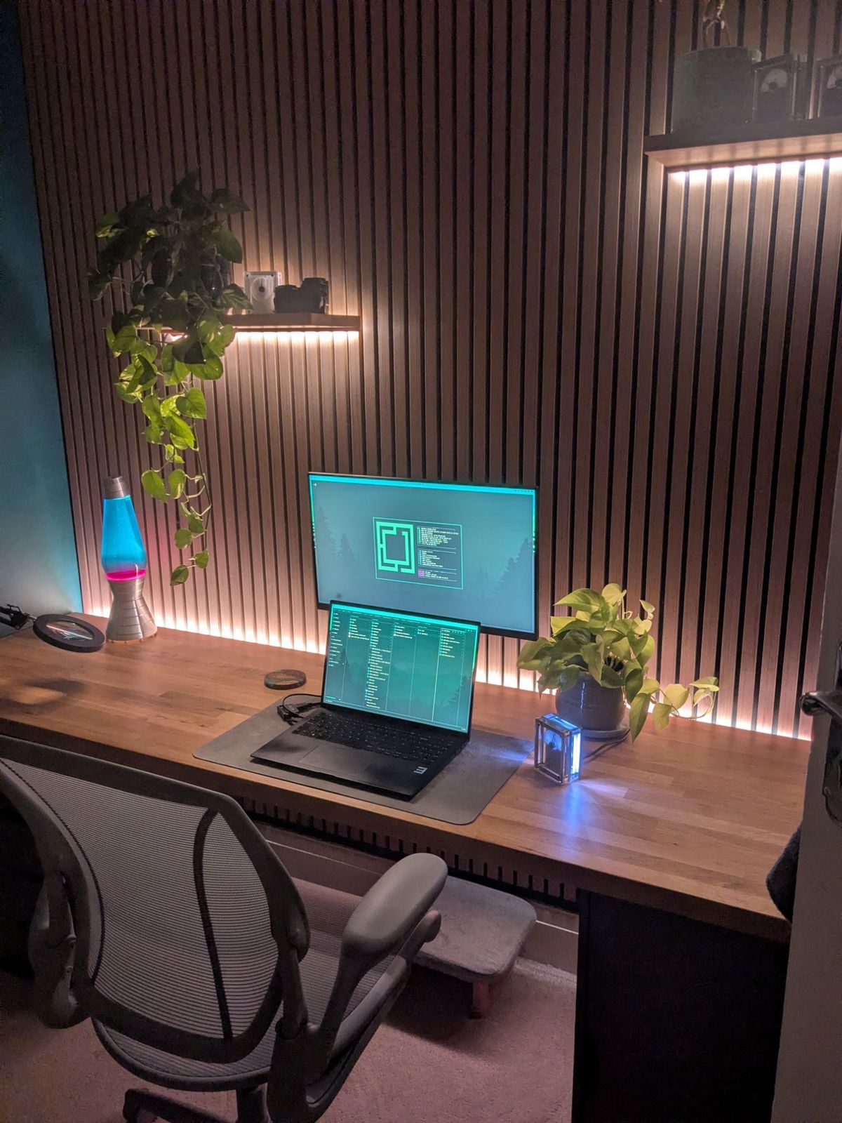 The office after dark, shot upright: the backlit slat wall, the lava lamp and the laptop and monitor showing teal code.