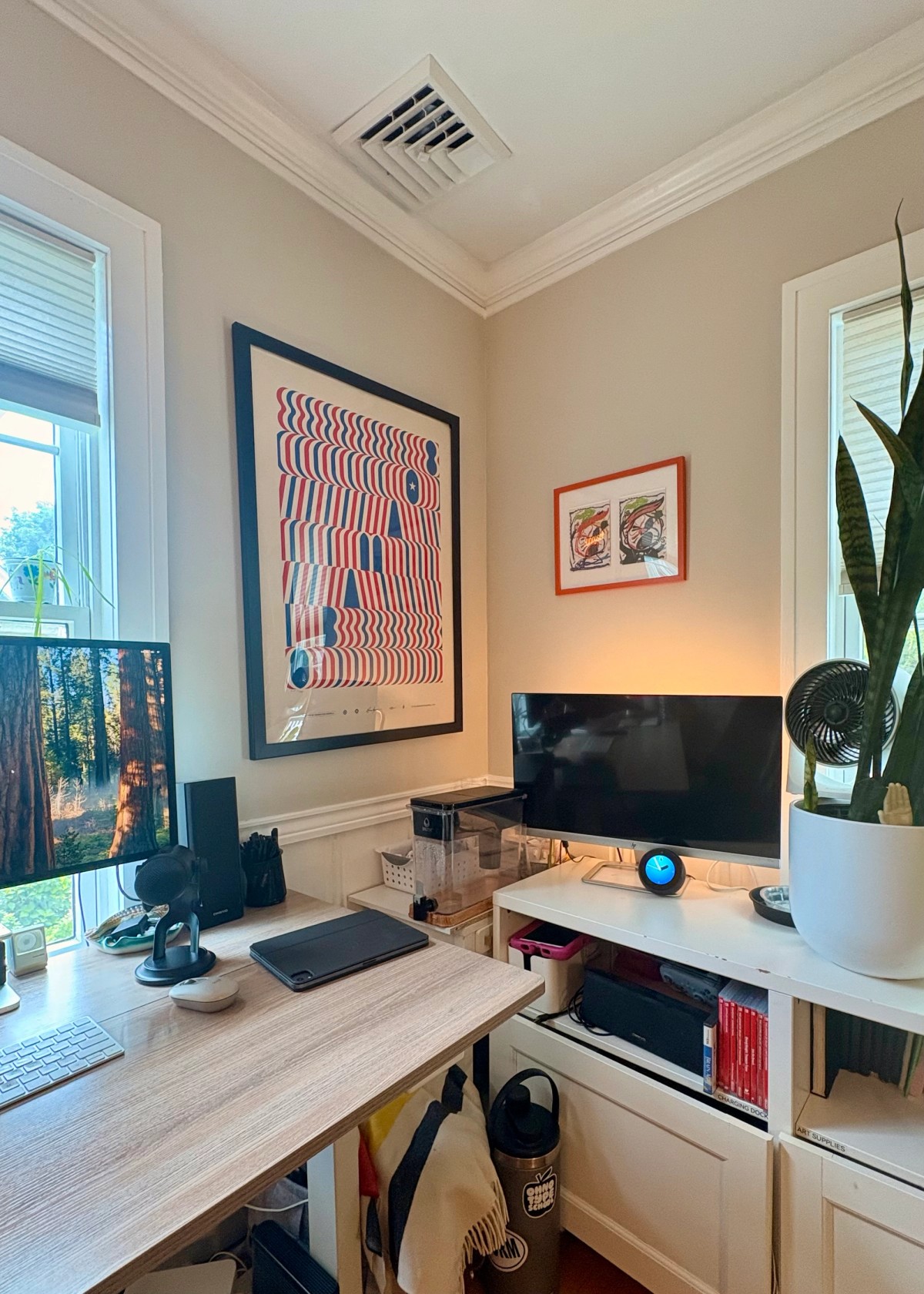 Zack Davenport's standing desk desk by a window with an Apple keyboard, an iPad, a microphone, and a monitor showing a redwood forest, beneath a large red, white, and blue wavy op-art poster.