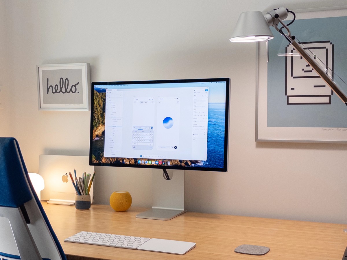 A minimal light-wood desk with an Apple Studio Display showing a Figma file of ChatGPT mobile screens. A MacBook Pro sits closed in a vertical stand beside a ceramic cup of colored pencils, a yellow HomePod mini, and a glowing IKEA TOKABO lamp. Two framed Susan Kare prints hang on the wall — a hand-lettered "hello." and the pixel Happy Mac — with an Artemide Tolomeo lamp arcing in from the right.