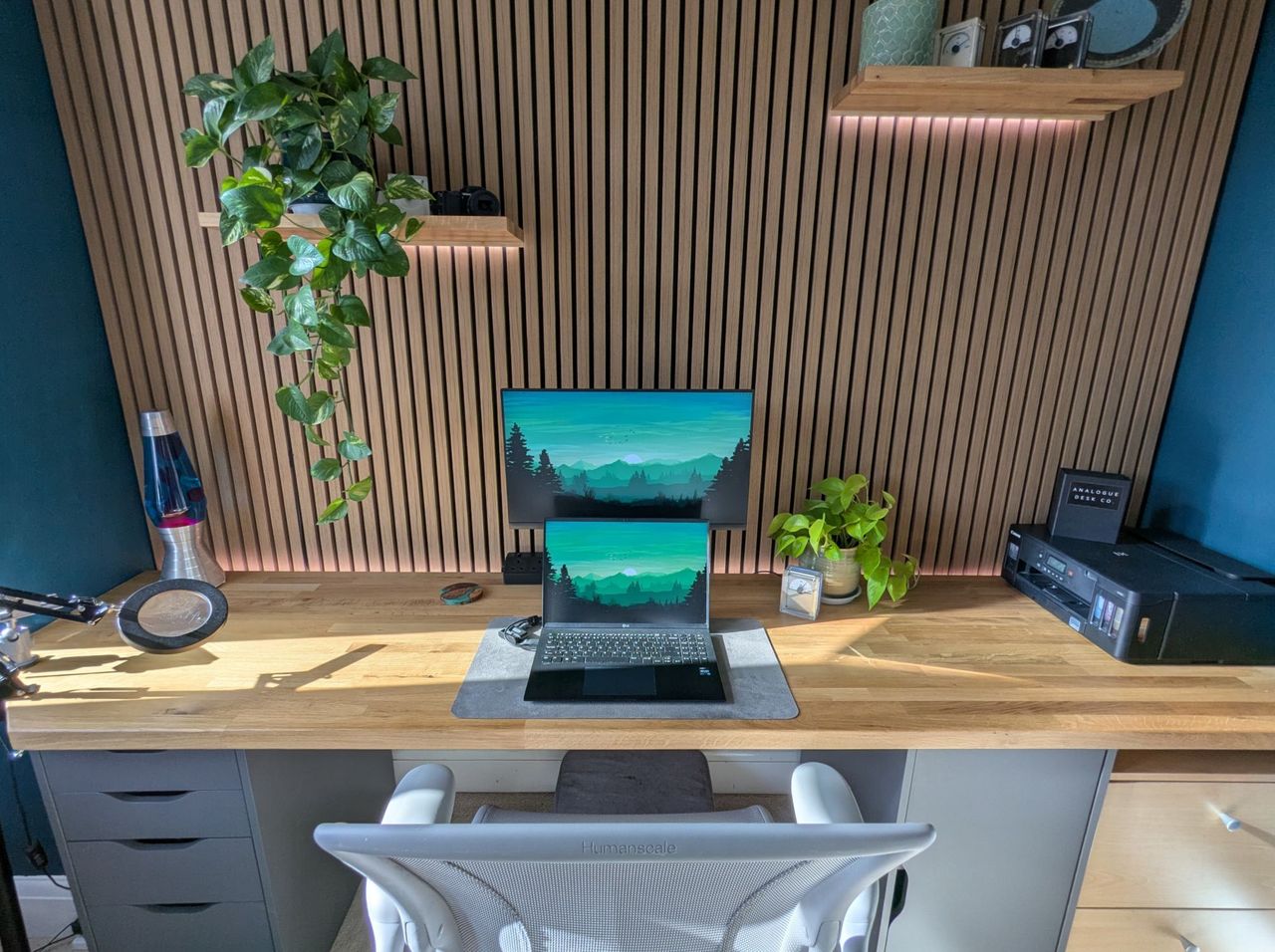 A wider head-on view of the desk with the lava lamp at one end and a printer at the other, under the backlit slat wall.