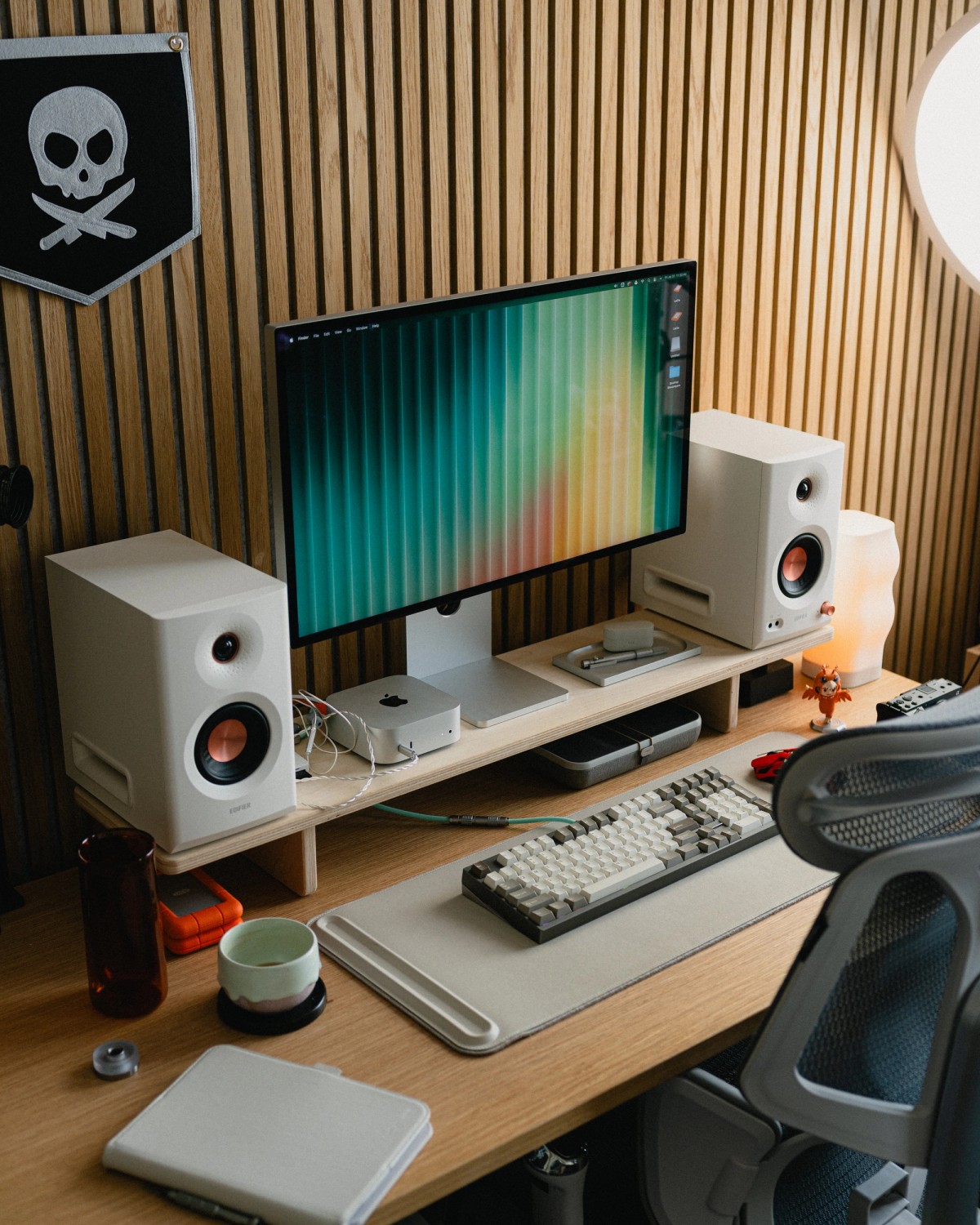 A wood-slat home office with white bookshelf speakers either side of a monitor showing a teal gradient, and a black skull pennant on the wall.