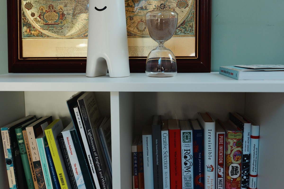 A white bookcase packed with books on design, typography and photography, with a smiling vase and an hourglass on the shelf above, in front of a framed antique world map.