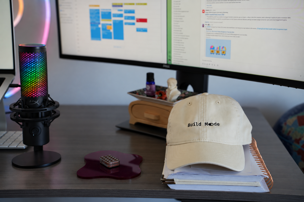 HyperX QuadCast microphone beside a Dell ultrawide monitor, with a Build Mode cap resting on a stack of notebooks