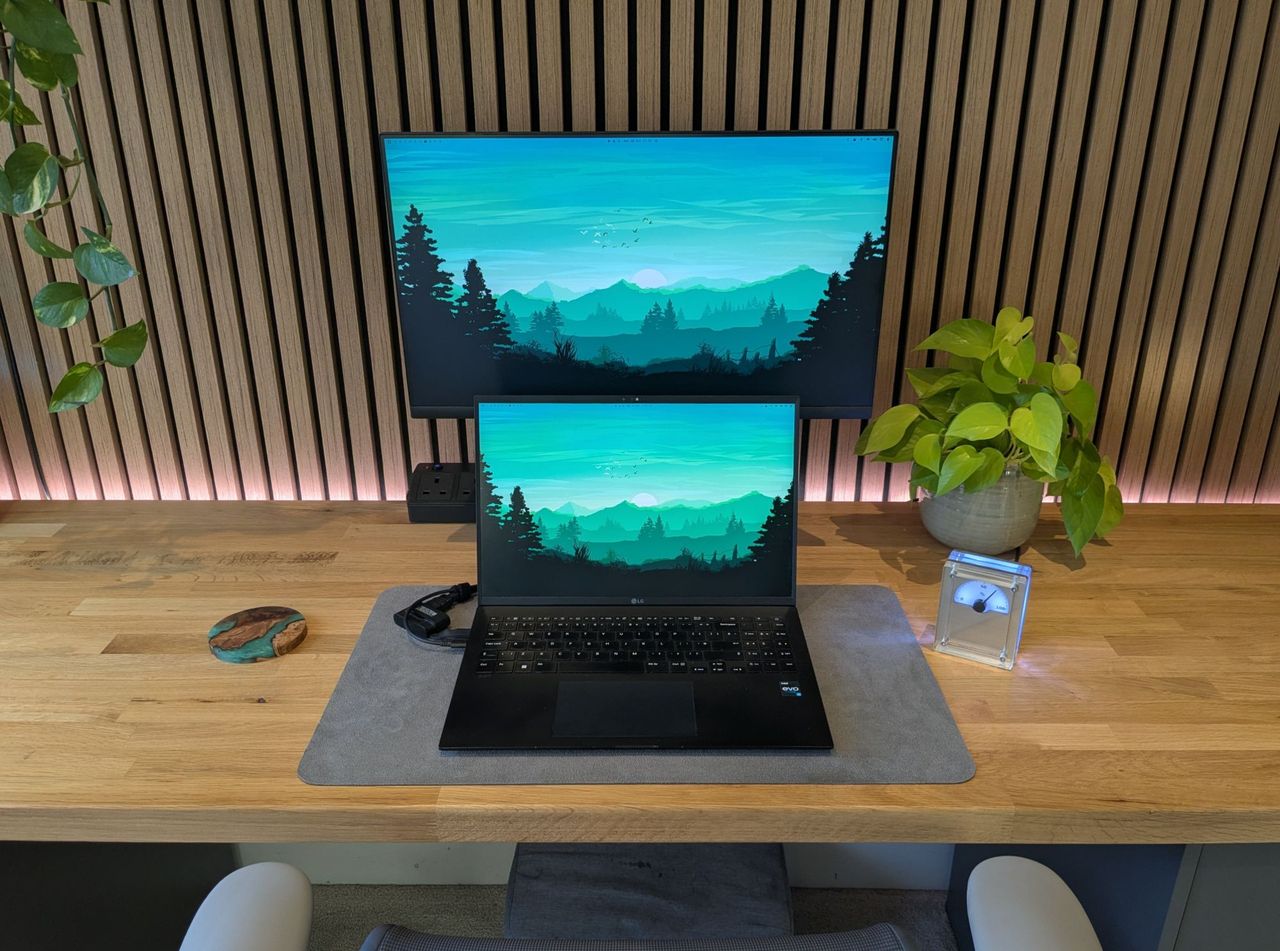 The desk head-on: a Dell monitor and an LG Gram laptop both showing a teal mountain wallpaper, with a plant and the desk gauge beside them.