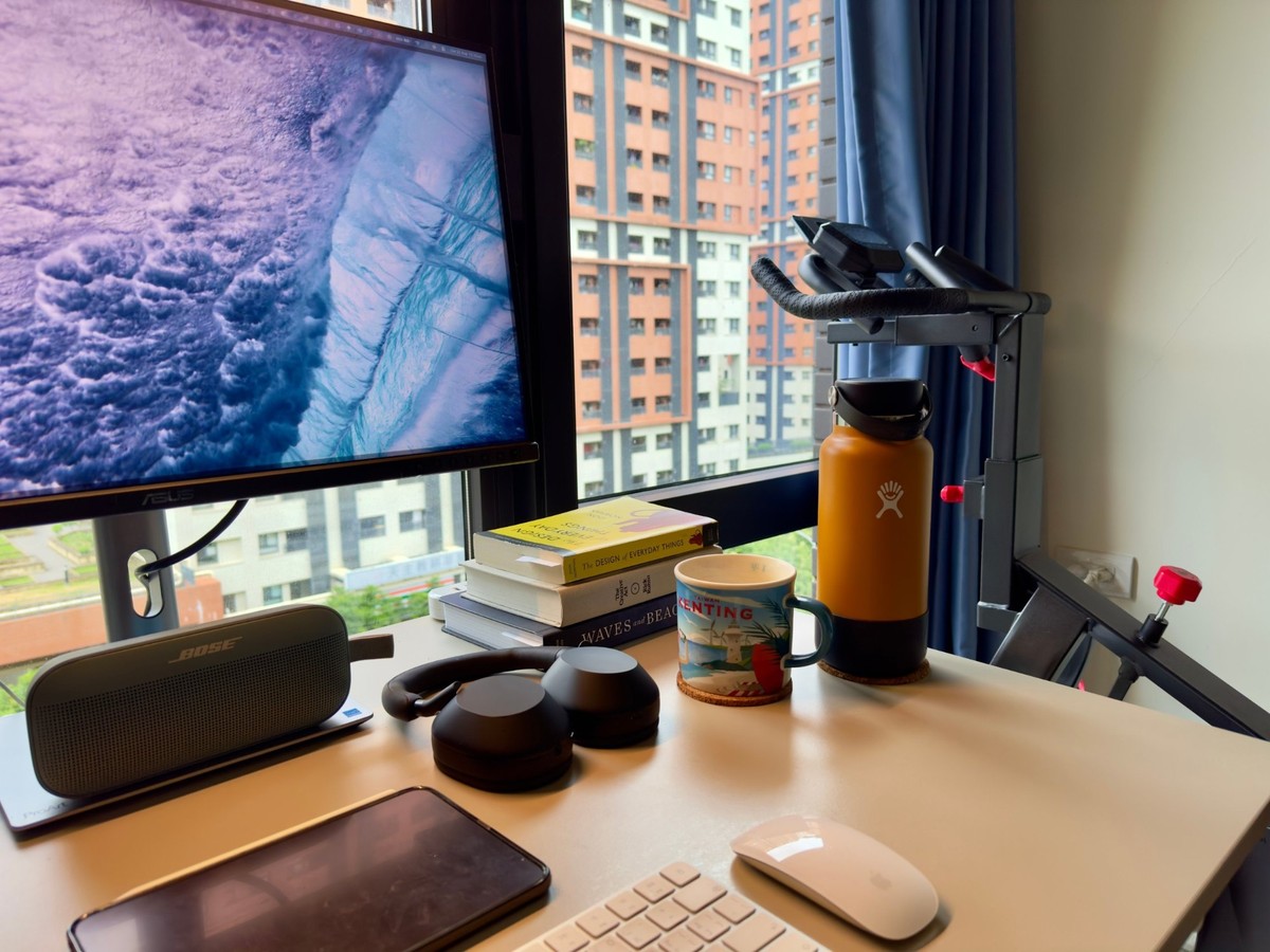 A close view across the desk: a stack of books including The Design of Everyday Things and The Creative Act, a Taiwan Kenting mug on a cork coaster, an orange Hydro Flask and the handlebars of an exercise bike by the curtain.