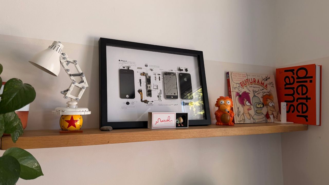 A floating oak shelf holding a Lego Luxo Jr. lamp, a framed iPhone 4 teardown, a Marcel the Shell box, a Simpsons monkey figure and a Dieter Rams book.