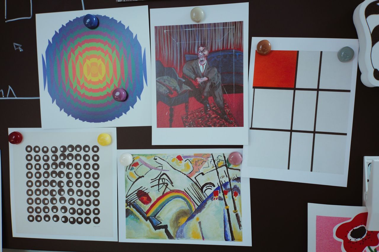 A close view of art postcards held to a dark board with colored magnets, including an op-art spiral, a figurative painting and a grid composition.