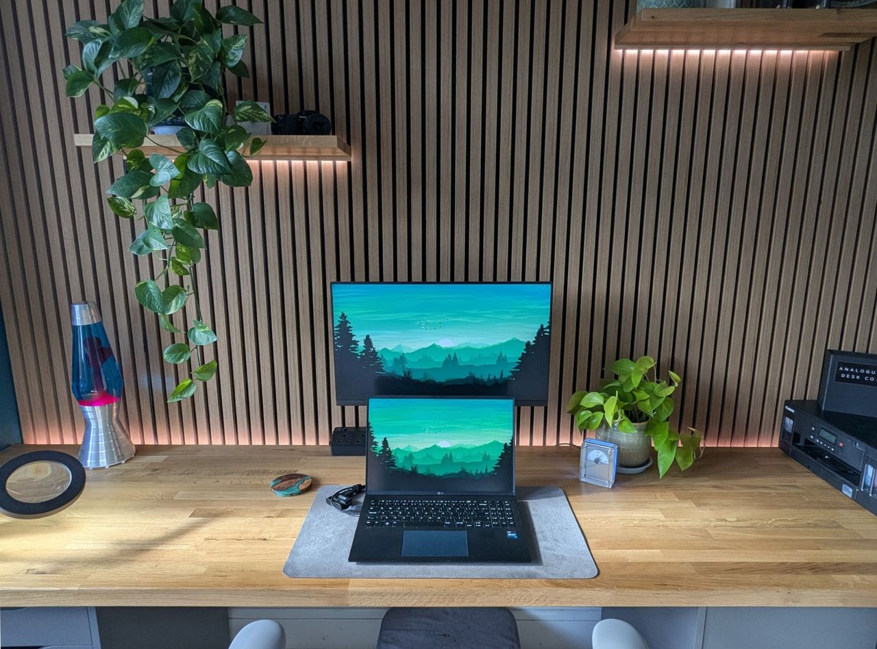 The oak slatted wall running the length of the desk, backlit in warm orange, with a lava lamp, trailing plants and a floating shelf.