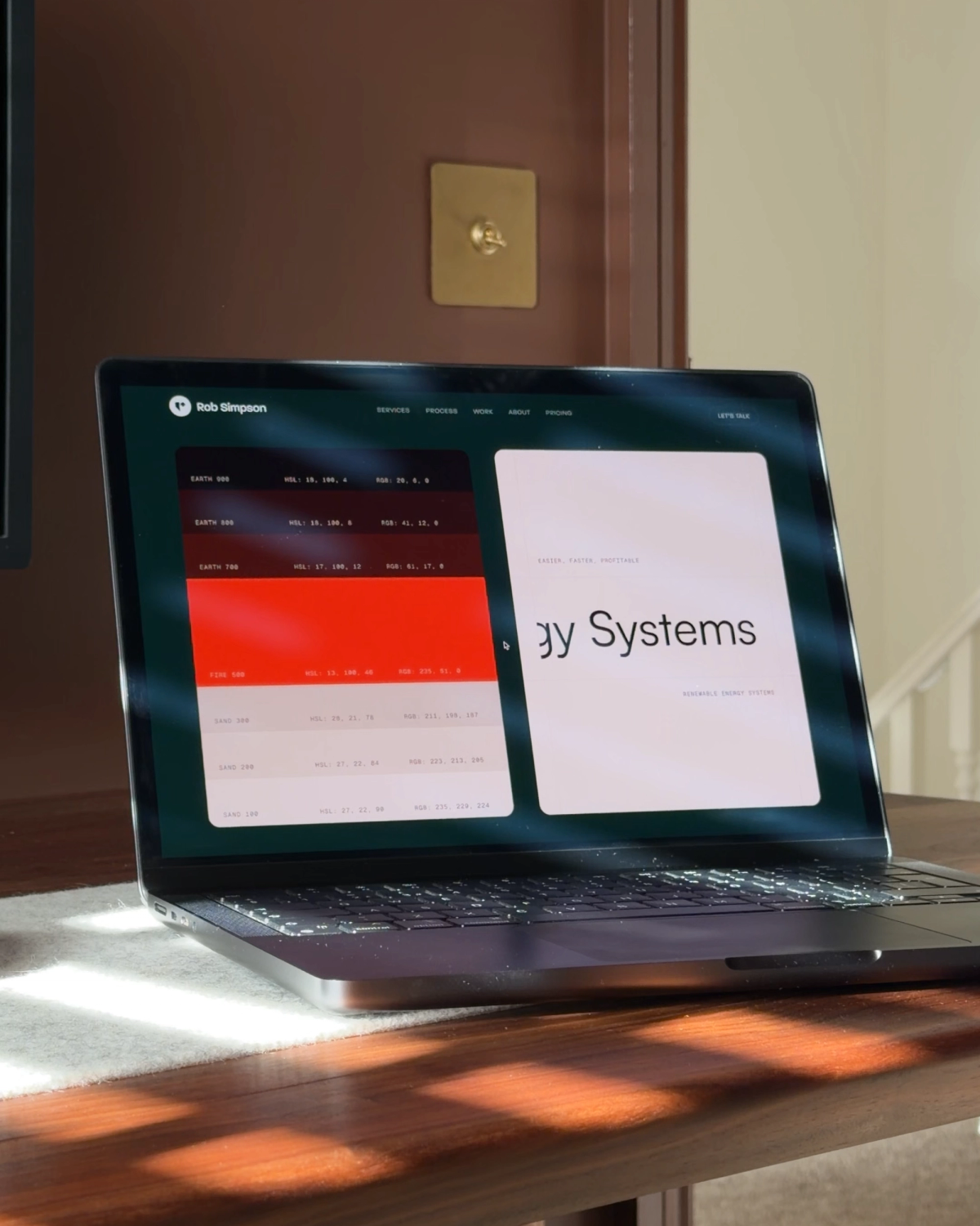 A MacBook Pro catching morning sun, showing a color palette of earth and fire tokens next to a client homepage, with a brass toggle switch on the wall behind.