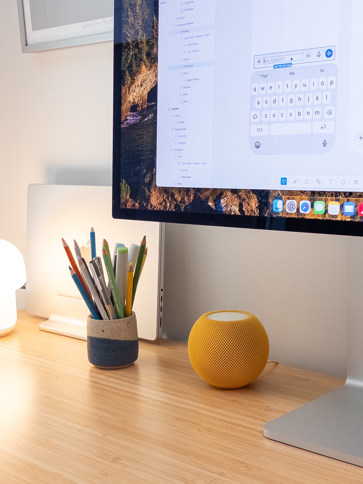 Close-up of the left side of Gavin Nelson's desk. A stoneware cup holds Prismacolor pencils and pens next to a yellow HomePod mini and the edge of a MacBook Pro in a vertical stand. Behind them, the Studio Display shows a Figma layer panel and an iOS keyboard mockup with an "Ask ChatGPT" composer field selected.