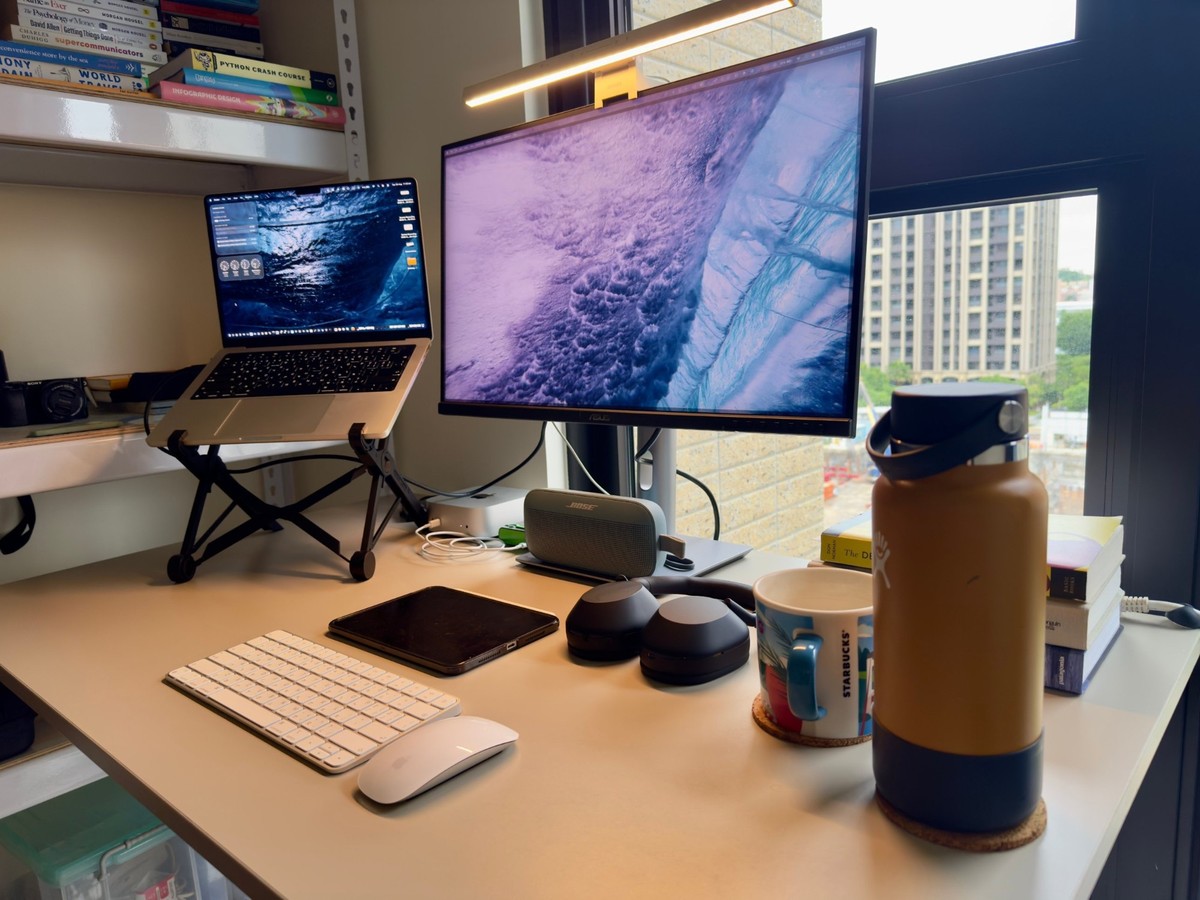 The desk seen from the right, with the Mac mini and a Bose speaker tucked under the monitor, an iPad beside the keyboard and a white bookshelf above the laptop.