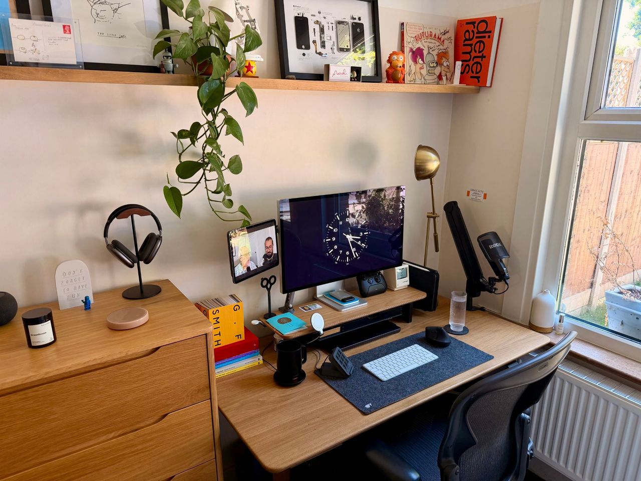 A light oak desk in the corner of a bedroom under a shelf of framed art, with an Apple Studio Display, a brass lamp and a microphone on an arm, and a mesh chair in front of the window.