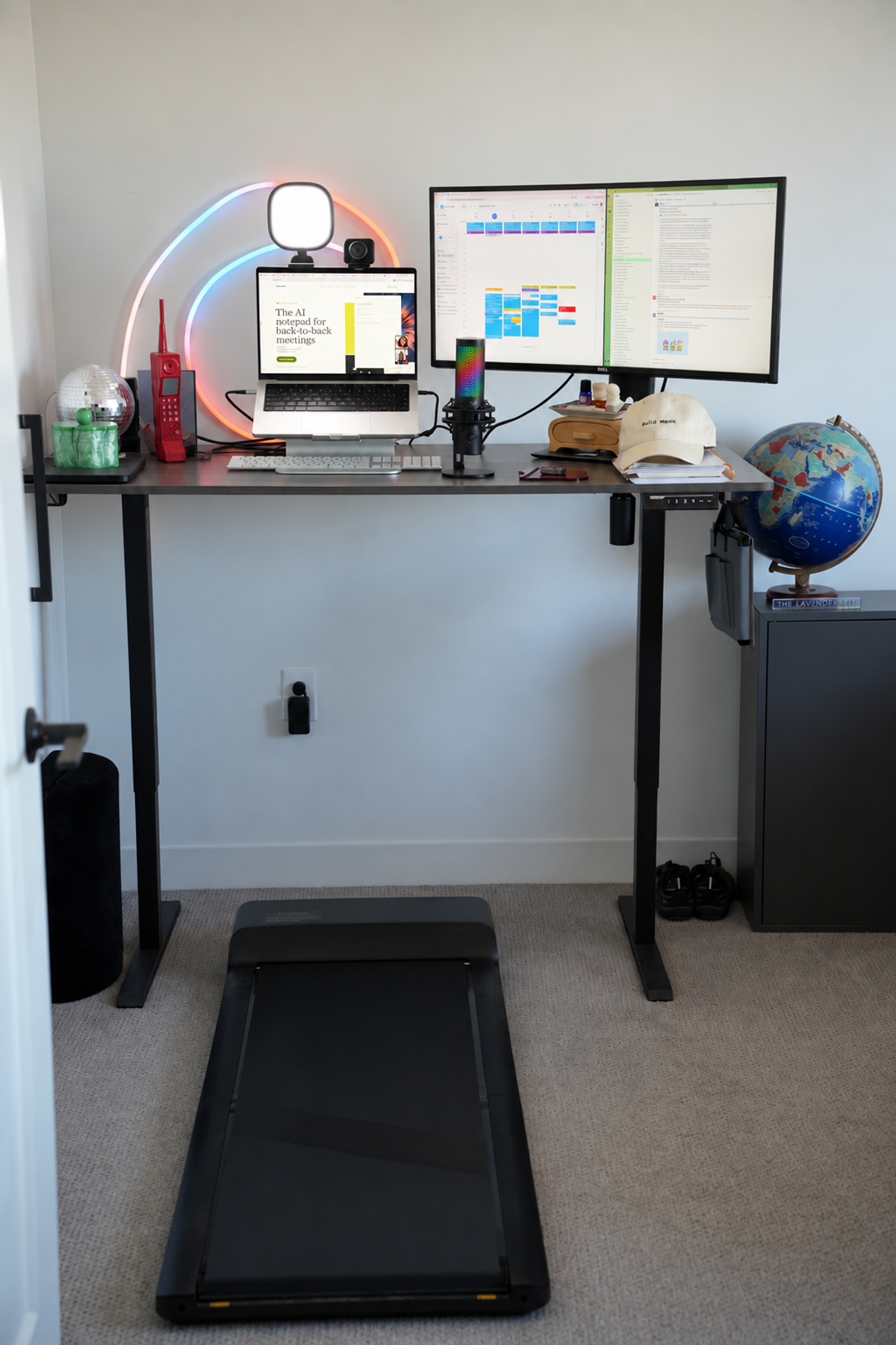 Compact home office with a black standing desk, a curved ultrawide monitor, a neon ring light on the wall and a foldable walking pad on the carpet below