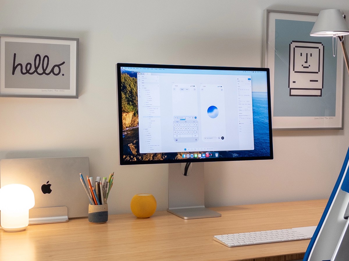The desk photographed from the left, with the blue Steelcase Gesture chair in the foreground. A Magic Keyboard and Magic Trackpad sit on the light wood surface below the Studio Display, lit by an Artemide Tolomeo lamp. Framed Susan Kare prints hang on the wall behind.