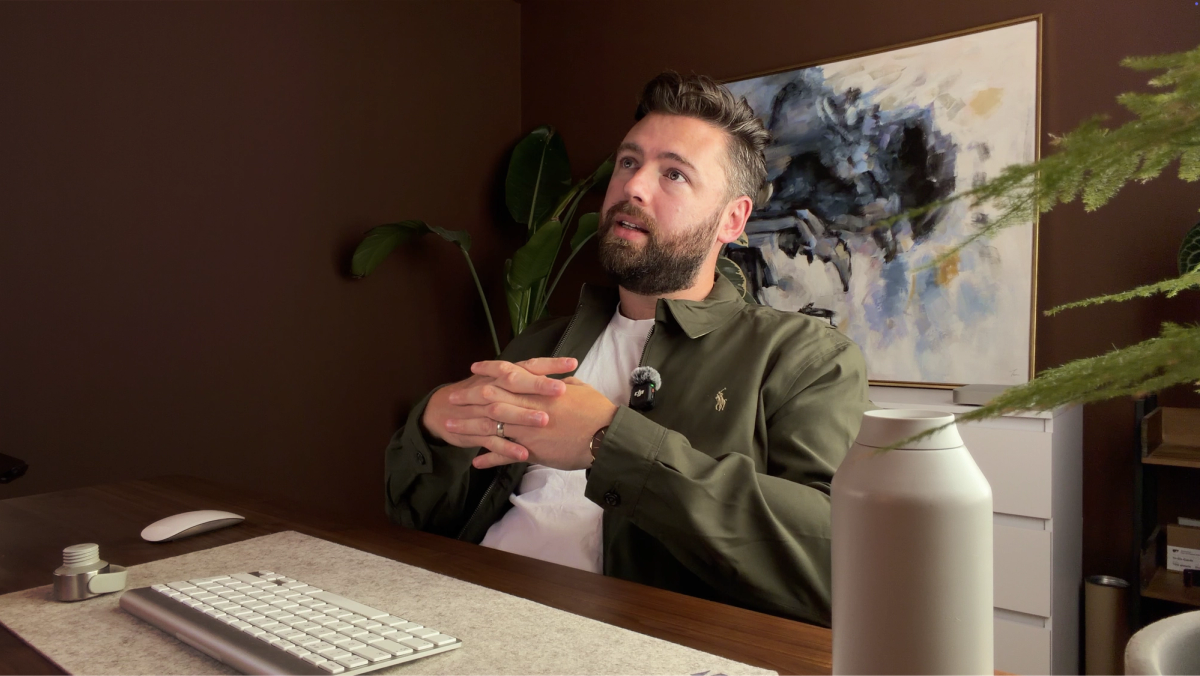 Rob at his desk mid-sentence, in a green jacket with a lapel mic, a painting and a large plant behind him.