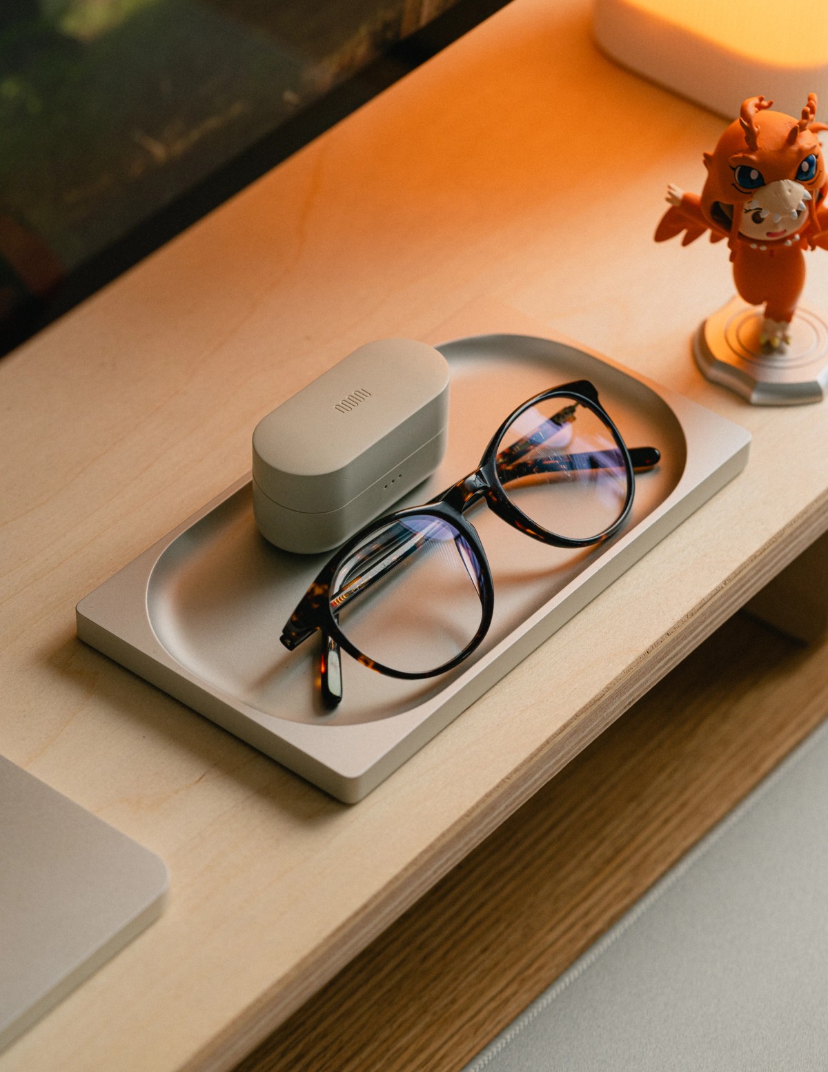 A pale stone tray holding tortoiseshell glasses and a small earbuds case on a plywood shelf.