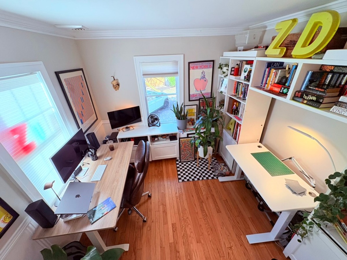 Overhead view of Zack Davenport's New York home office, with a light-wood desk holding dual monitors and a MacBook, a white standing desk with a green cutting mat, and book-filled white shelves topped by large yellow "ZD" marquee letters.