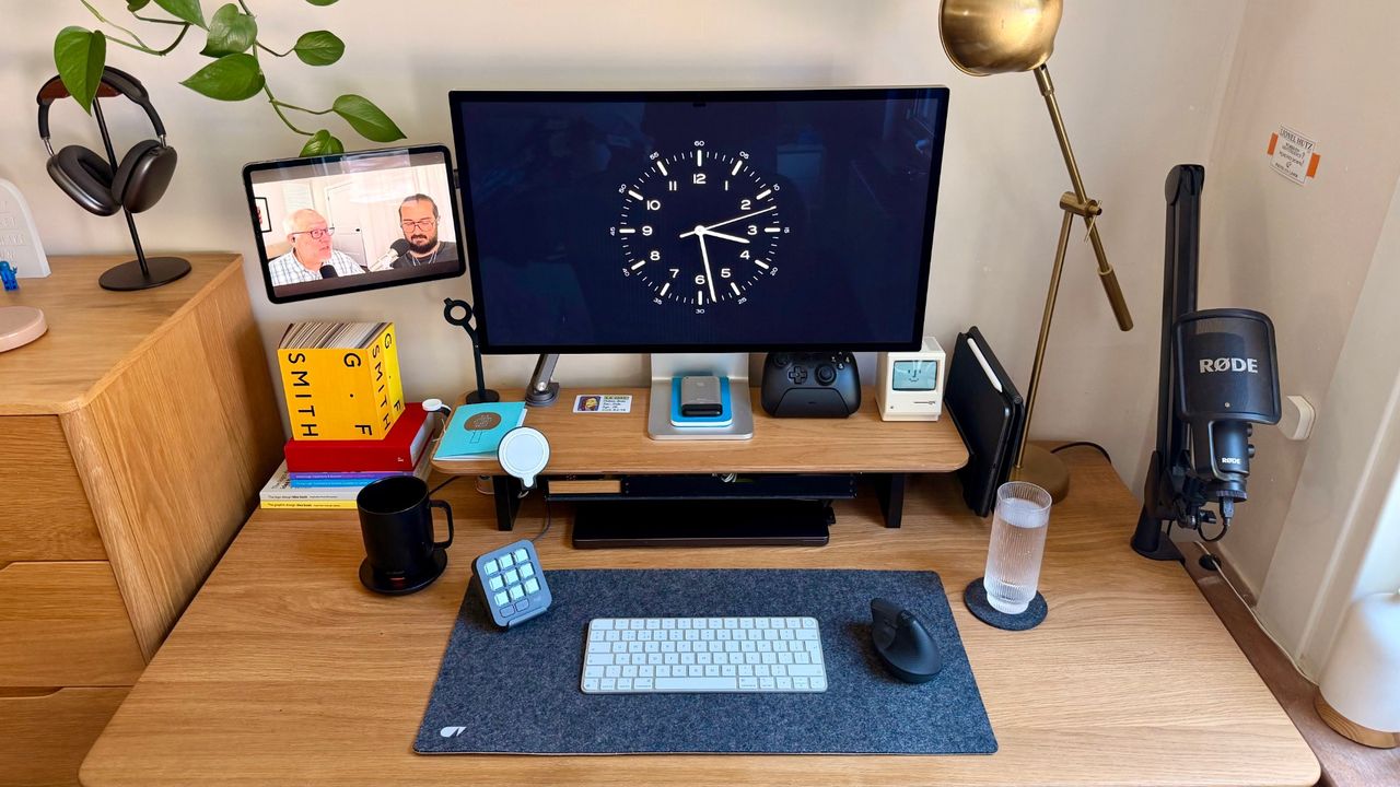 The desk head-on: an Apple Studio Display showing a clock face, an iPad playing a podcast, AirPods Max on a stand, a RØDE microphone on an arm and a Logitech MX Creative Console beside the keyboard.
