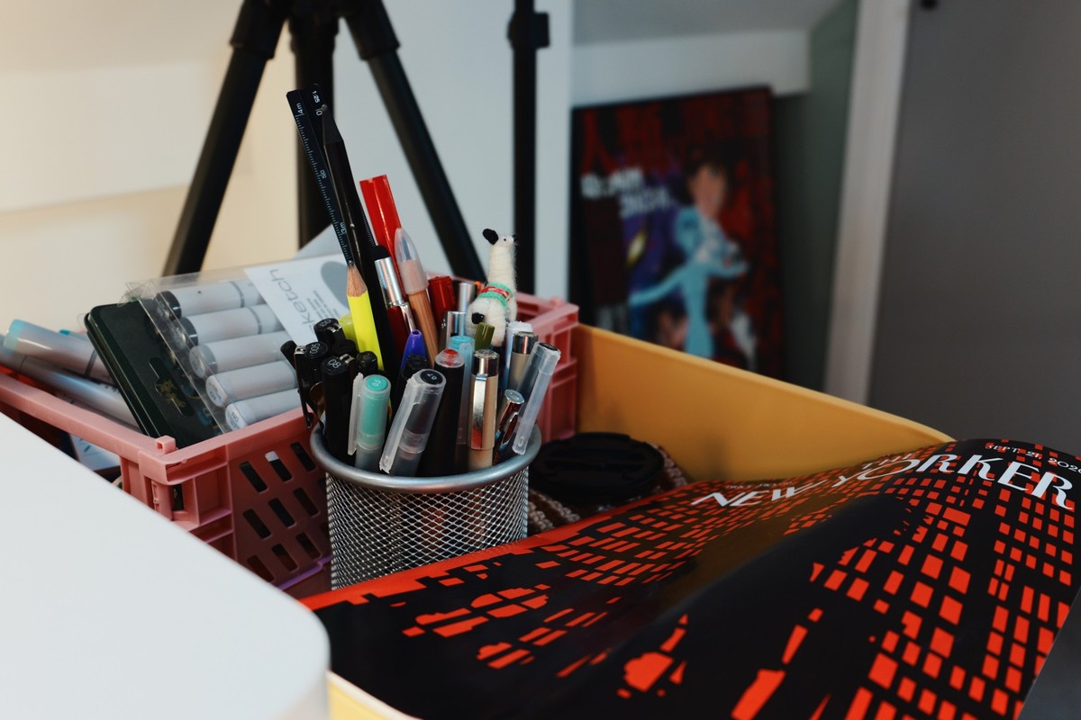 Pens, pencils and colored markers in a wire pot and pink crates on a yellow trolley, beside a rolled New Yorker cover.