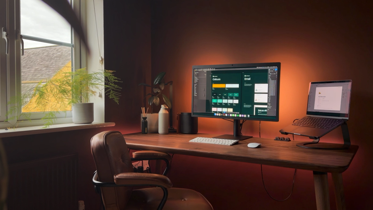 A walnut desk under a window, with an LG monitor showing a Figma file, a MacBook Pro on a stand and a brown leather chair, lit warm against a dark brown wall.