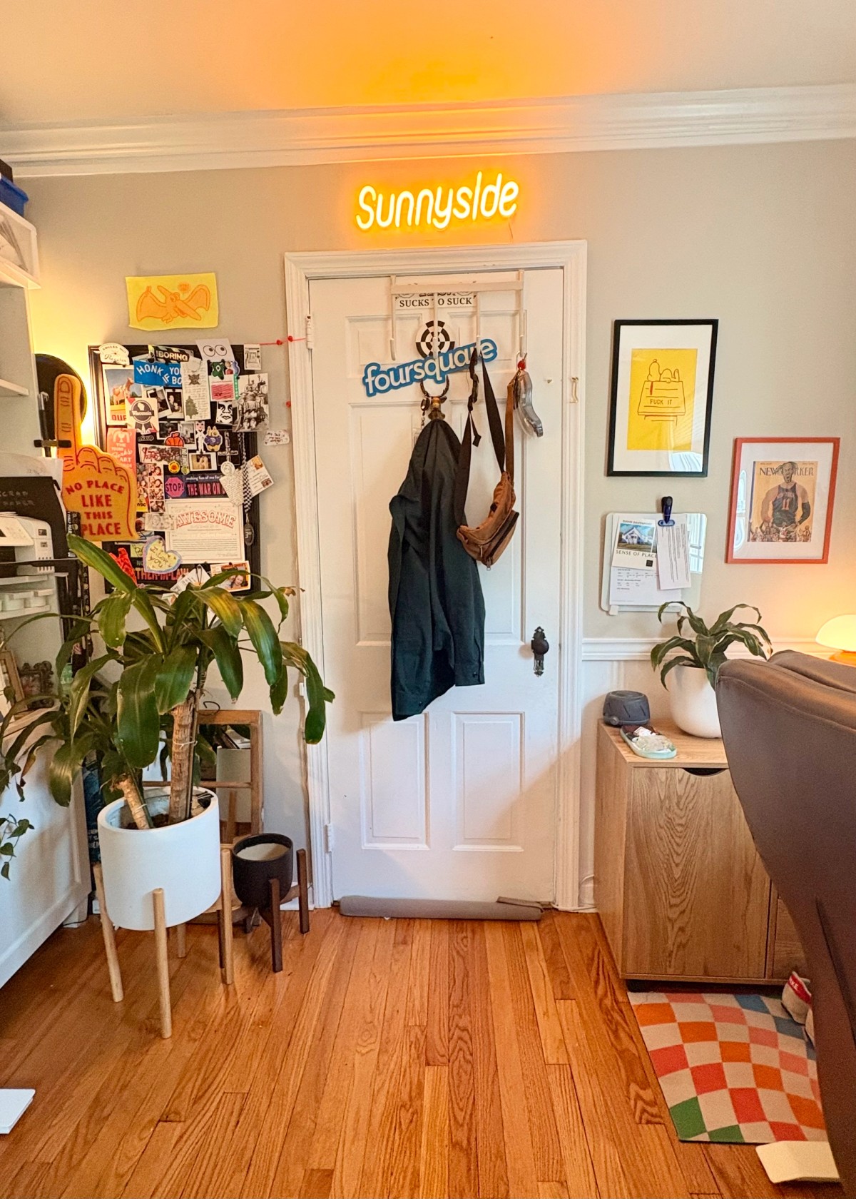 A white door topped by a glowing yellow "Sunnyside" neon sign, flanked by a sticker-covered vision board, a tall dracaena in a mid-century planter, and a leather desk chair.