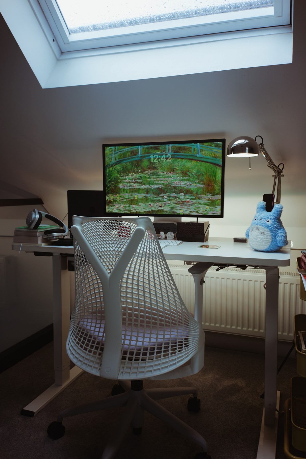 The office under a rain-flecked skylight, with a white Herman Miller Sayl chair pulled up to a white standing desk and the Studio Display lit against the wall.