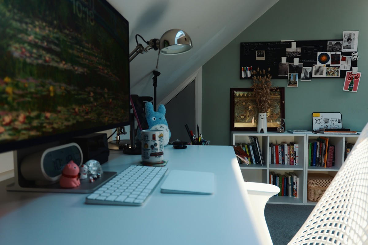 The desk seen from behind the chair, looking across to a green wall with a pinboard of photographs and prints above a white bookcase.