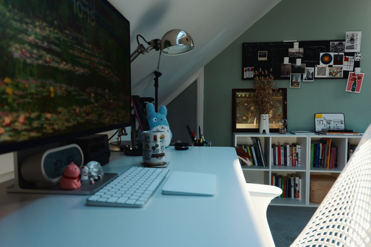 The desk seen from behind the chair, looking across to a green wall with a pinboard of photographs and prints above a white bookcase.