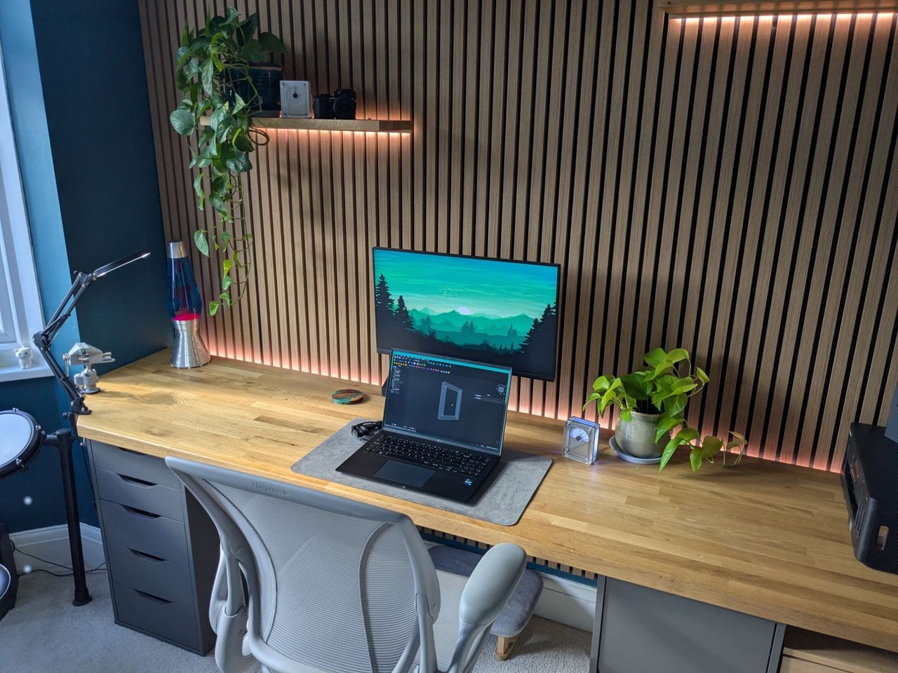 A long oak desk against a backlit slatted oak wall, with a monitor and laptop, a lava lamp, trailing plants and an electronic drum kit by the bay window.