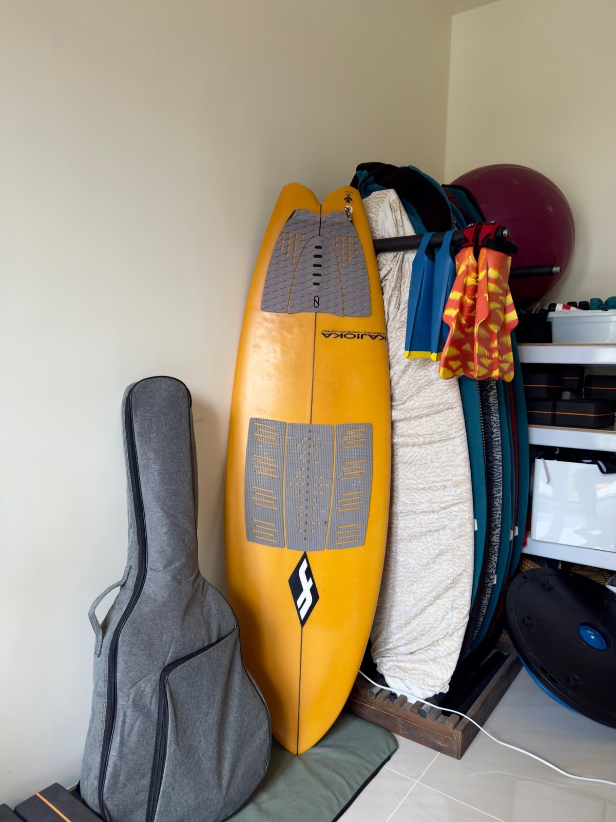 An orange surfboard leaning against the wall beside a guitar case, with fins, wetsuits and more boards on a rack behind it.