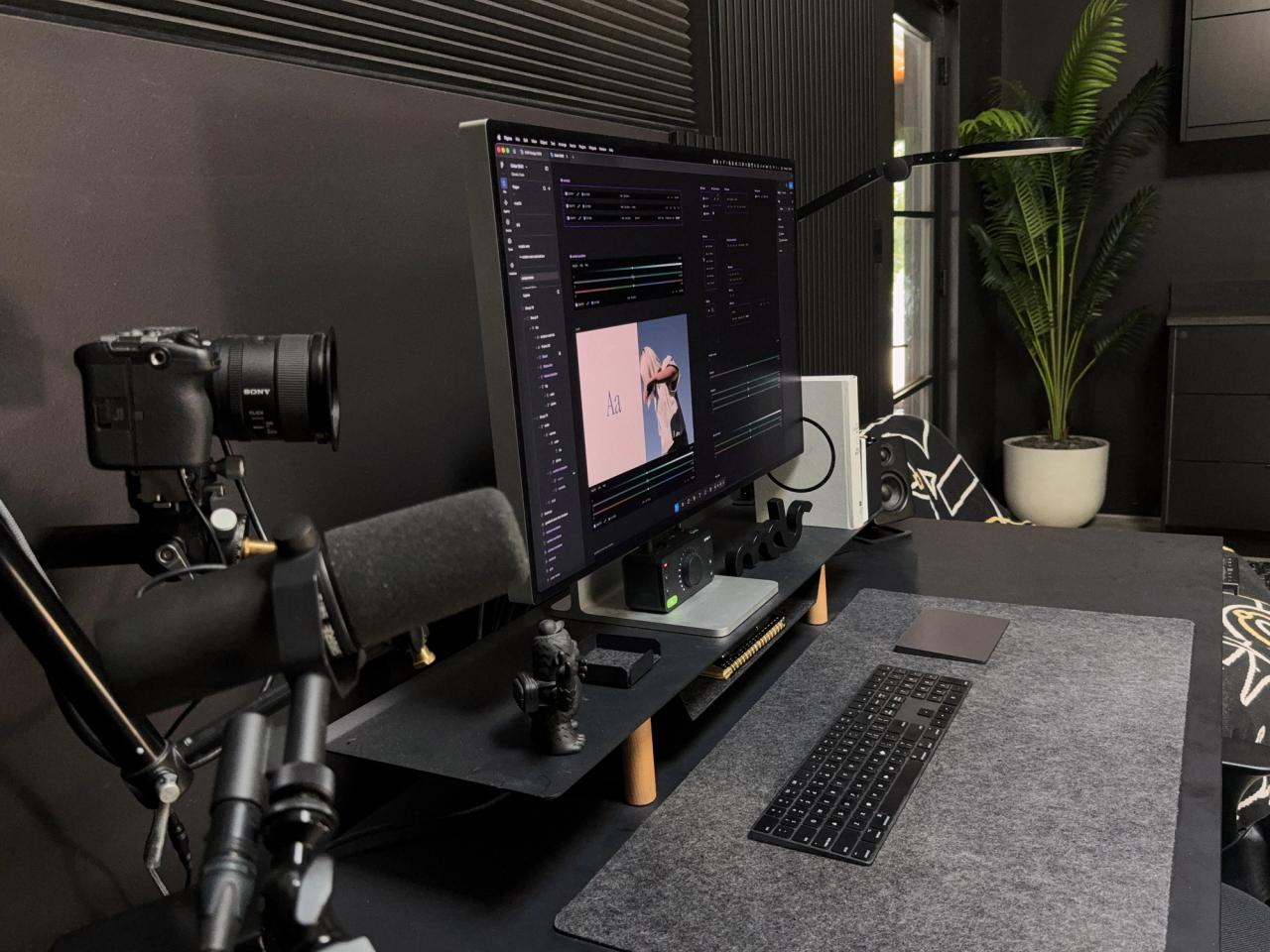 Side view of a black desk with a camera on an arm, a microphone, an Apple Pro Display XDR and a keyboard on a felt desk mat, with a palm in the corner