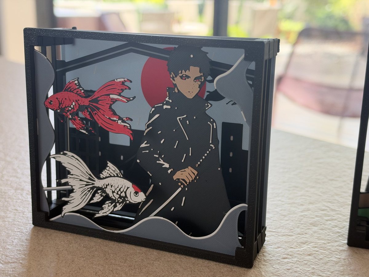 A paper theater seen at an angle, showing the gaps between its stacked panels: a swordsman in a long coat in front of a red sun and city buildings, with a red goldfish and a white goldfish swimming over grey waves in the foreground.
