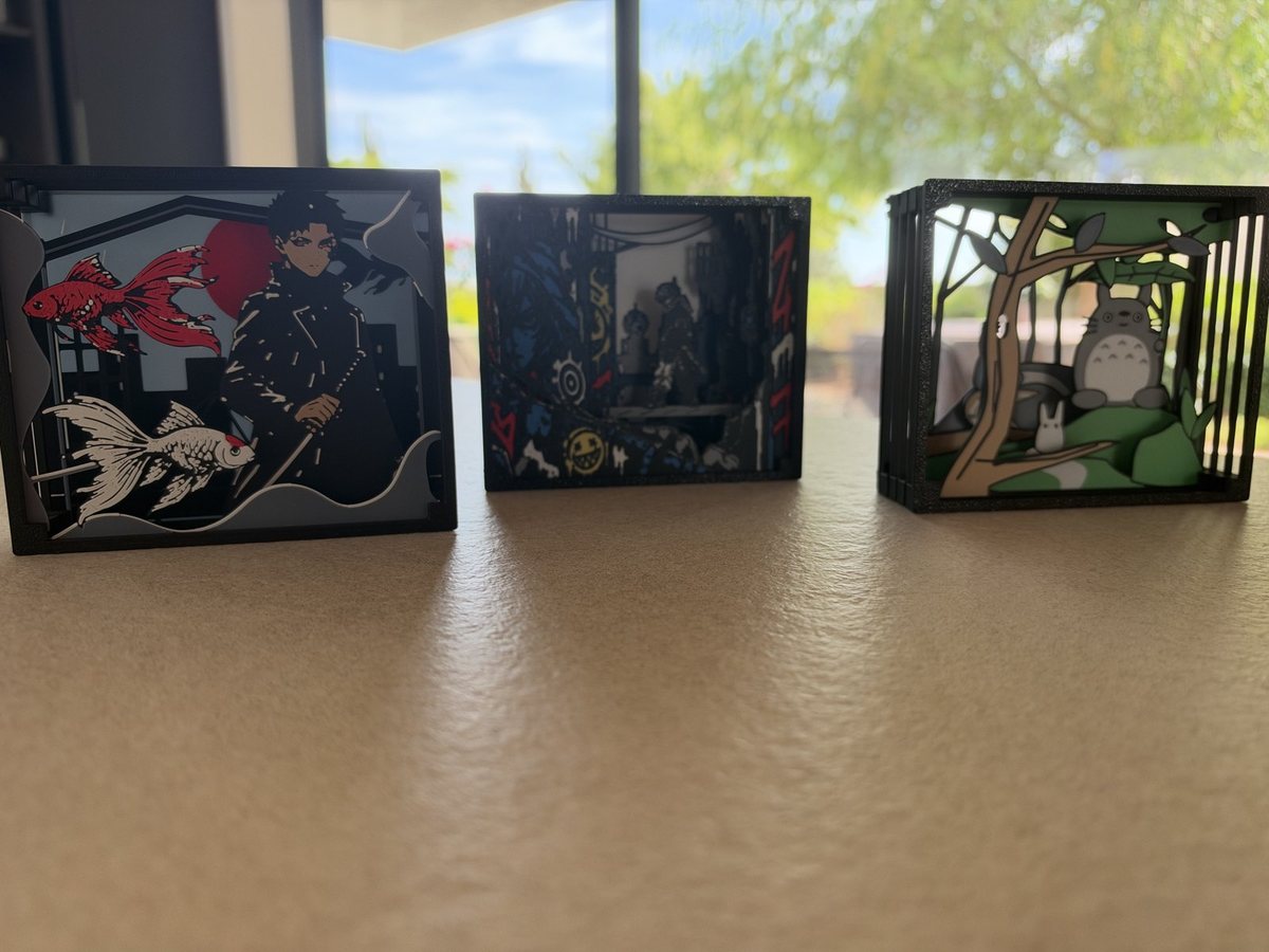 Three small black-framed layered paper theaters on a table in front of a window: a swordsman in a long coat with red and white goldfish, a dark graffiti-covered city alley, and a green forest scene with large round grey forest creatures beneath a tree.