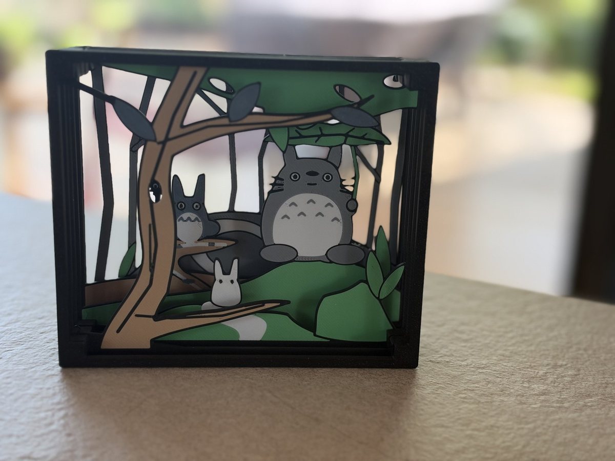 A paper theater of a forest scene: a brown tree trunk and dark leaves frame large and small grey forest creatures sitting on green hills, with pale sky showing through the vertical trees behind.