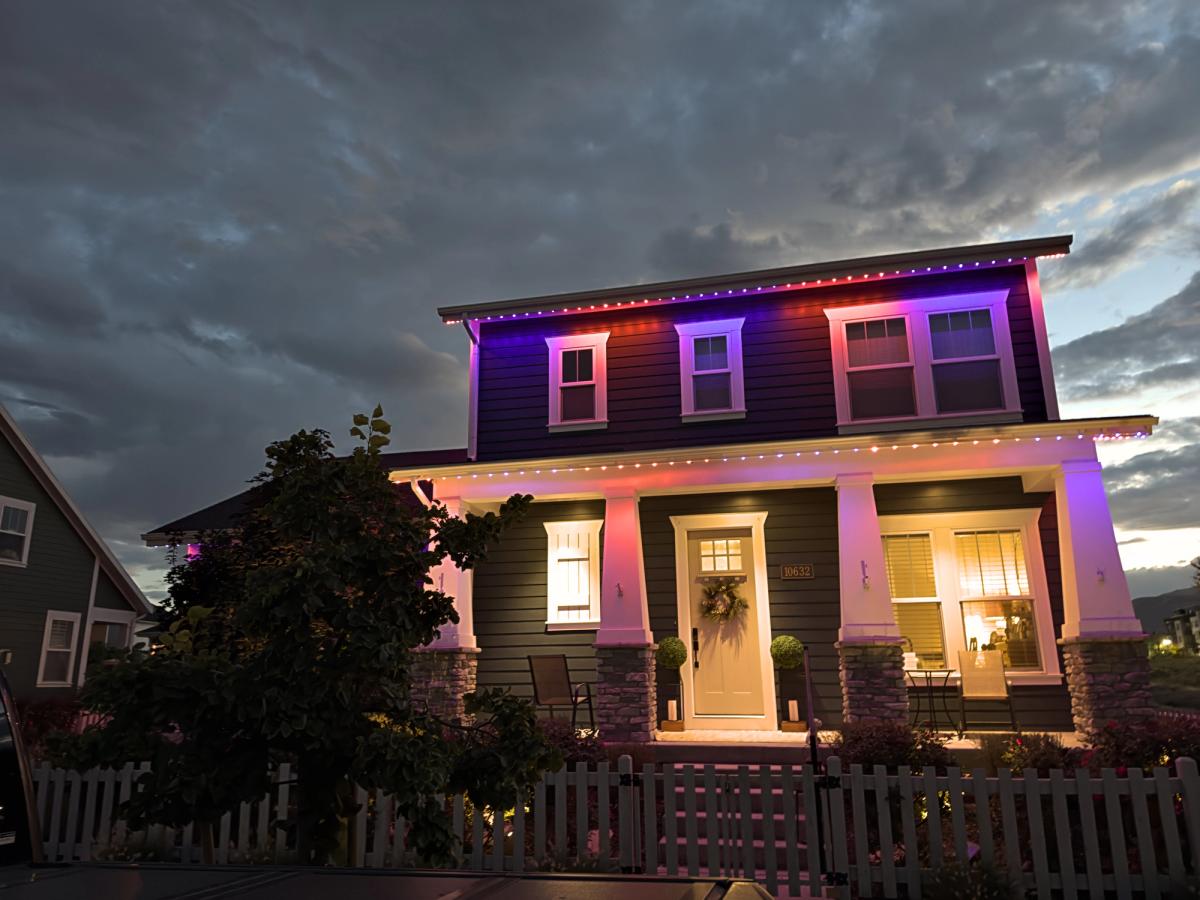 Two-story home with permanent outdoor LED lights glowing orange, purple, and warm white along the rooflines at dusk beneath a cloudy sky.