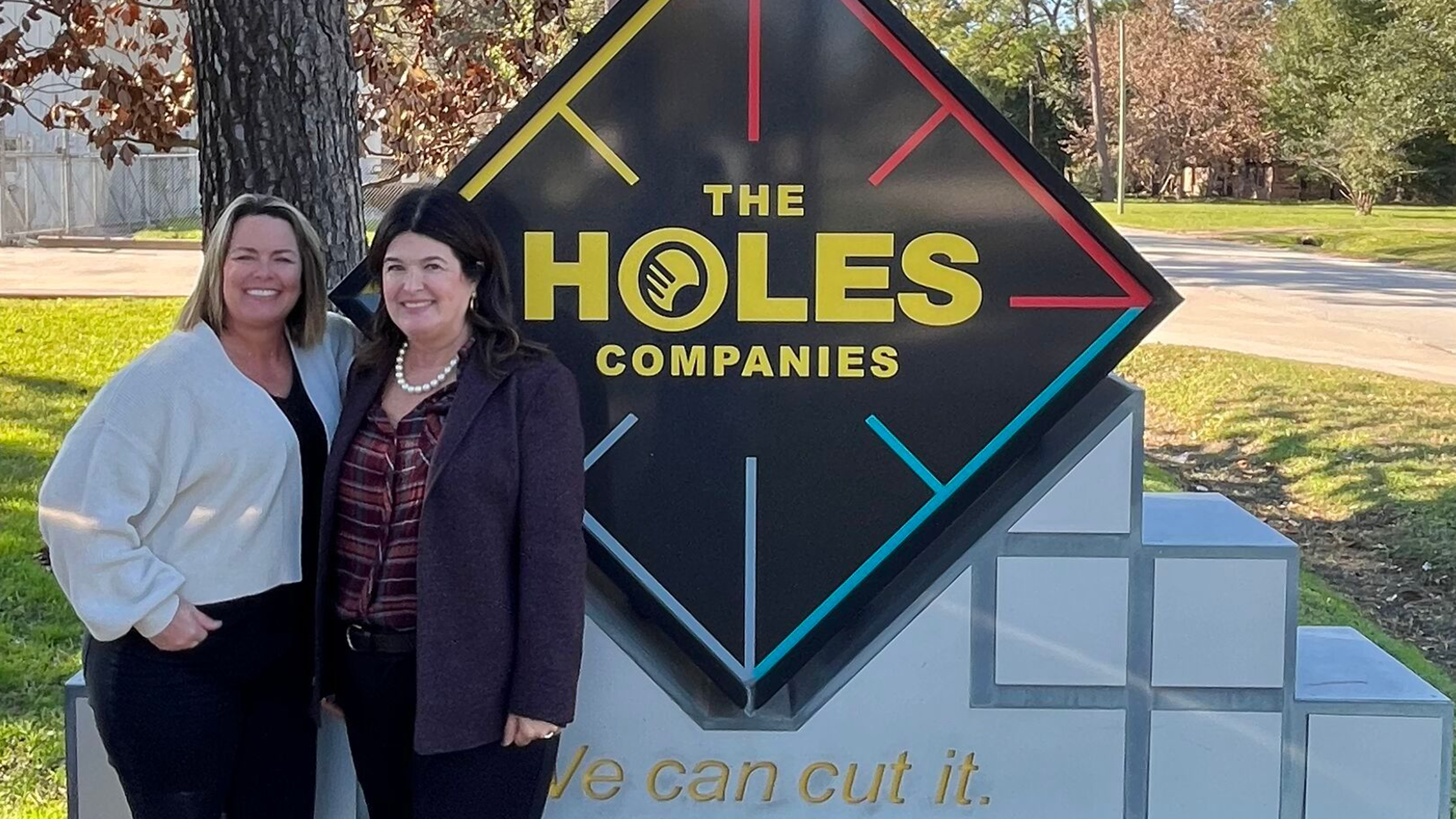 Darlene and Kelly stand in front of a sign that reads The Holes Company