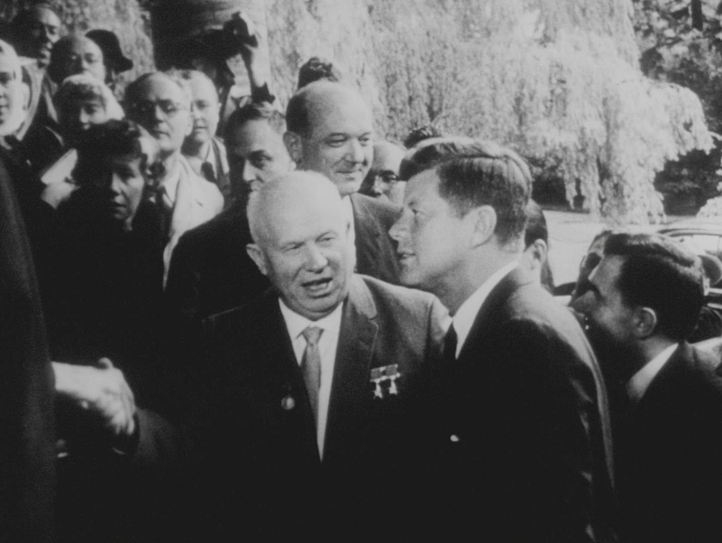 Black and white photograph of Nikita Khrushchev and John F. Kennedy next to each other during a state visit. Khrushchev has opened his mouth to speak, Kennedy can be seen from the side. Numerous people are standing in the background, a man is taking photographs.